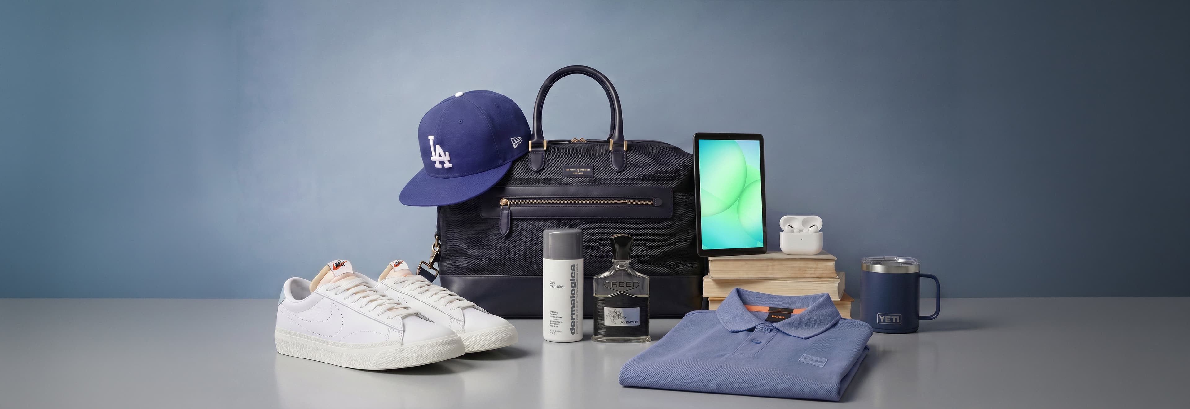 Collection of men's accessories and clothing on a table, including white trainers, a blue baseball cap, a black bag, a tablet, cologne, deodorant, wireless earbuds, a blue polo shirt, a mug, and books stacked nearby.