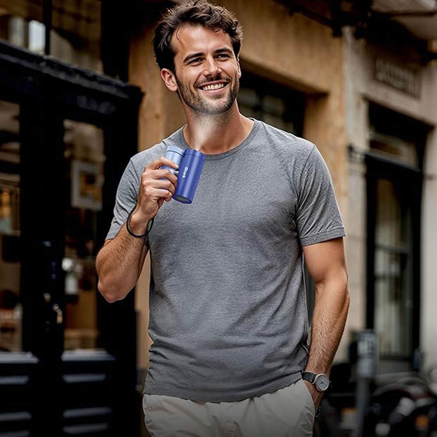 Man in grey t-shirt holding blue insulated bottle, standing in urban courtyard setting for summer essentials campaign