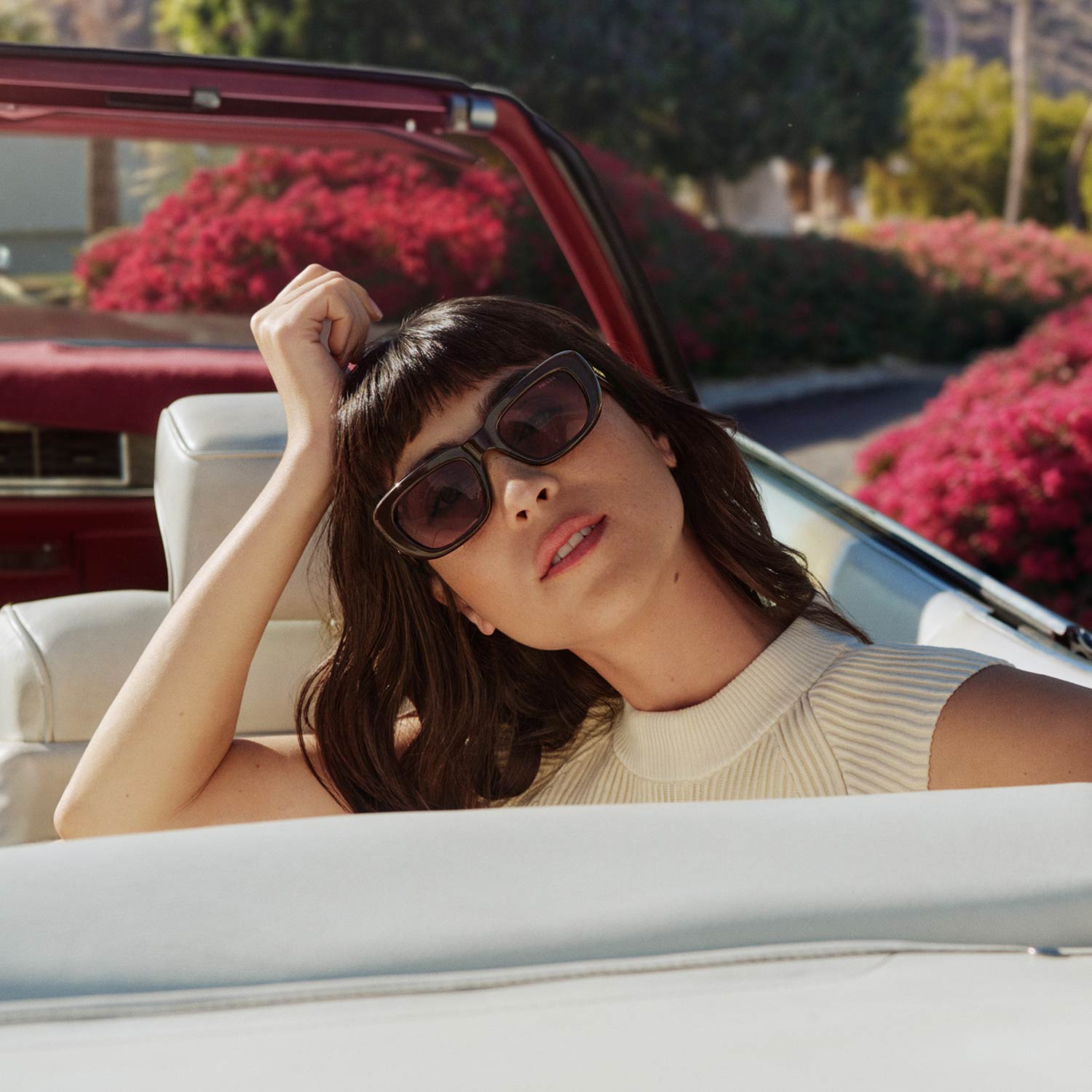 Woman wearing dark-framed sunglasses and cream ribbed top, relaxing in a convertible car with pink flowering plants in the background.