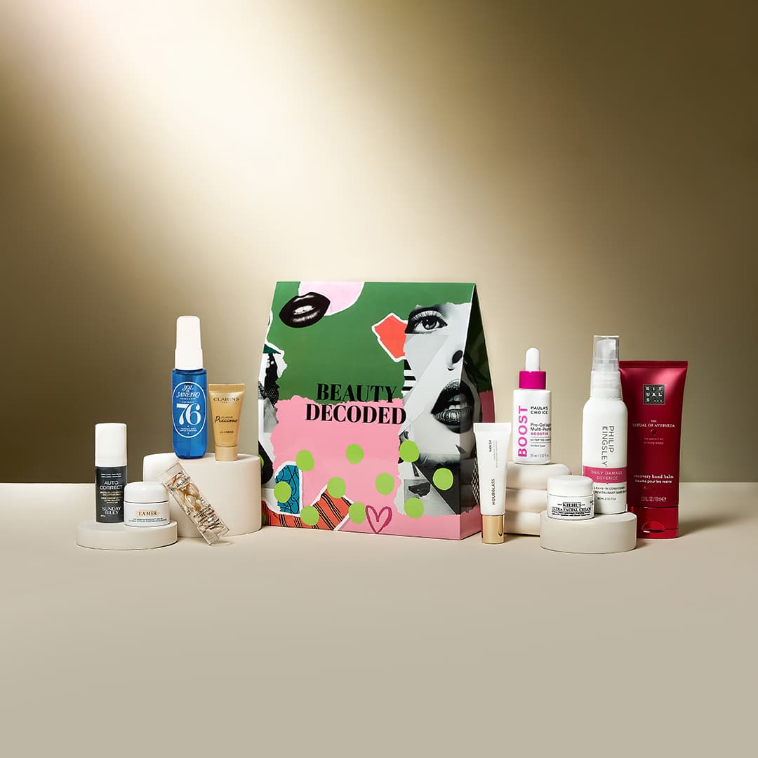A collection of beauty products arranged around a colourful box labelled 'Beauty Decoded'. Products include creams, sprays, and serums from various brands, displayed on a beige surface with a warm gradient background.