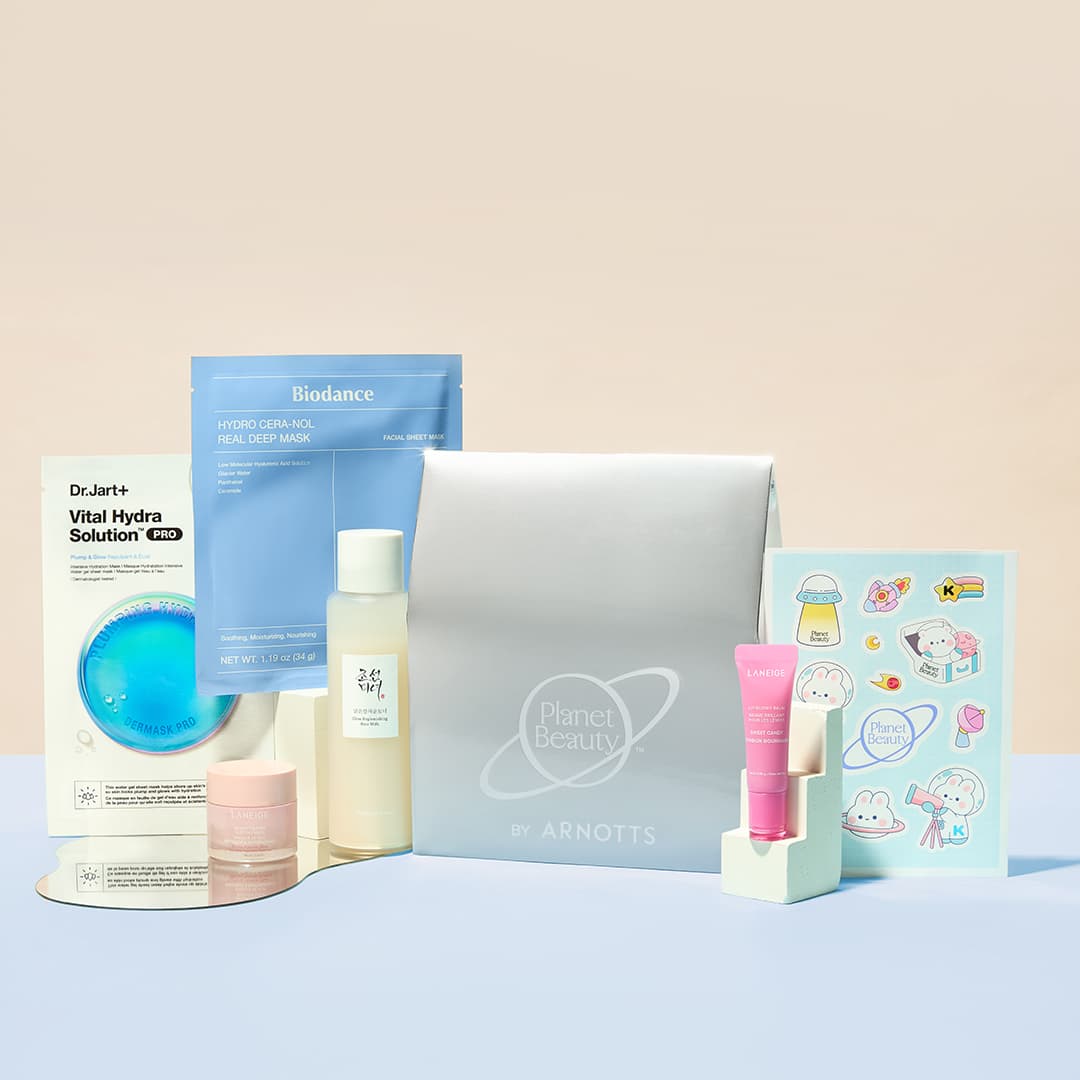A collection of beauty products arranged around a silver gift box with 'Planet Beauty' and 'ARNOTTS' written on it. Items include a Dr. Jart+ mask, Biodance facial mask, a bottle of skincare product, a pink tube of cream, and a sheet of cute stickers in pastel colours. The background is a soft gradient of beige and light blue.