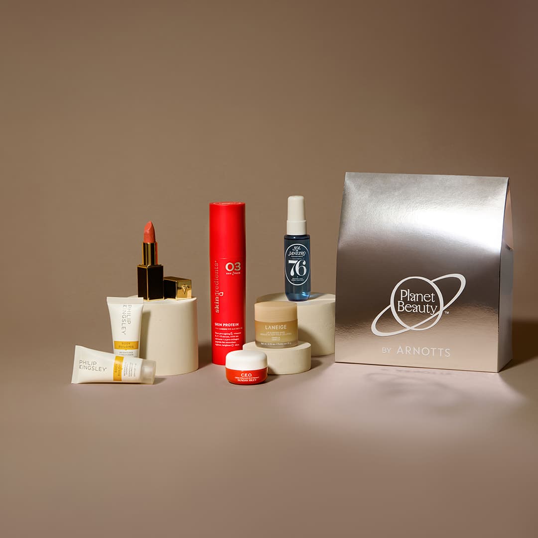 A collection of beauty products including a tube of cream, a lipstick, a red cylinder bottle, a spray bottle, and various small containers are arranged next to a silver gift bag with 'Planet Beauty' and 'Arnotts' printed on it. The items are displayed on cylindrical stands against a neutral background.