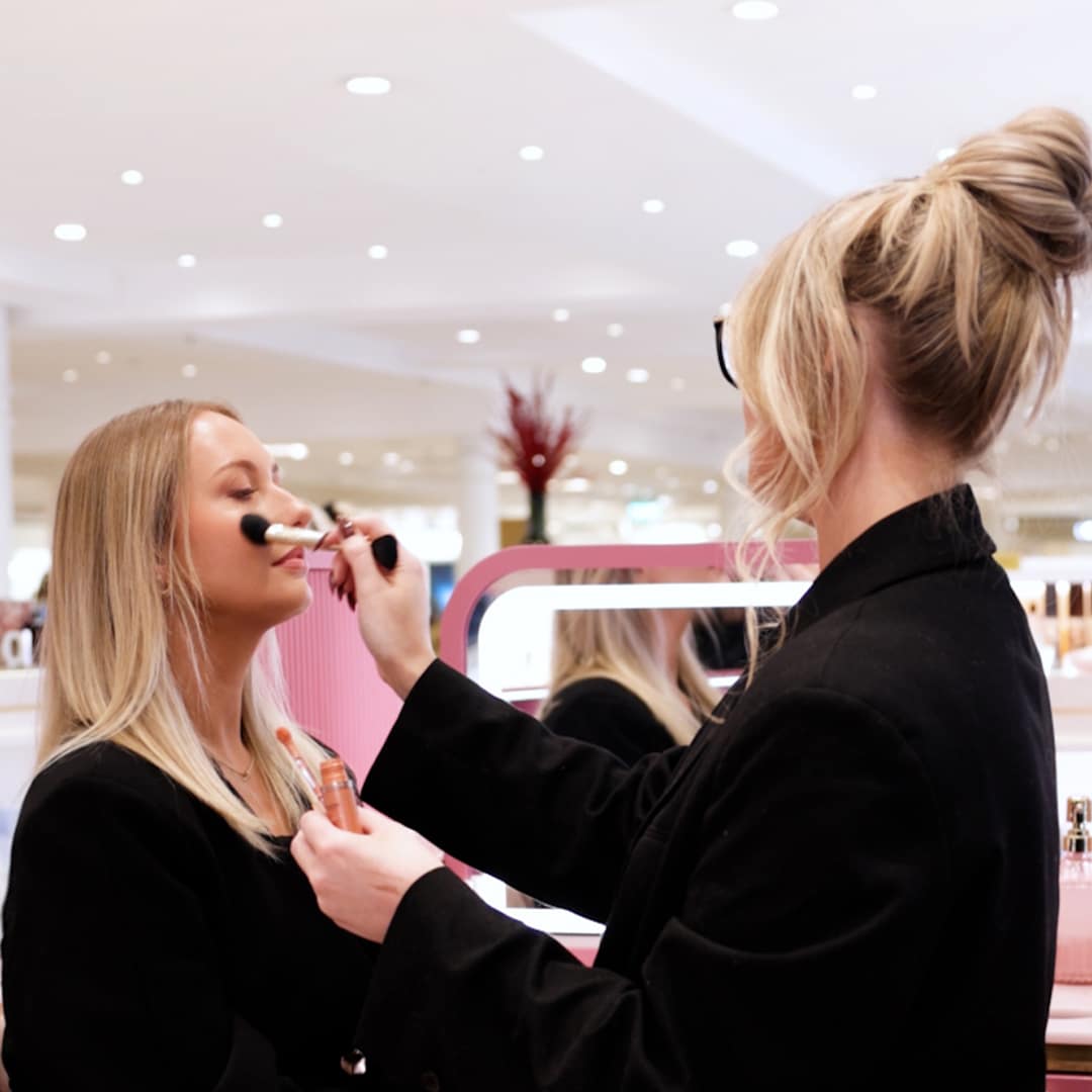 A make-up artist applies blush to a woman's face in a cosmetics store. The woman, with long blonde hair, is seated, while the artist holds a brush in one hand and a blush palette in the other. The store background features various beauty products and soft lighting.