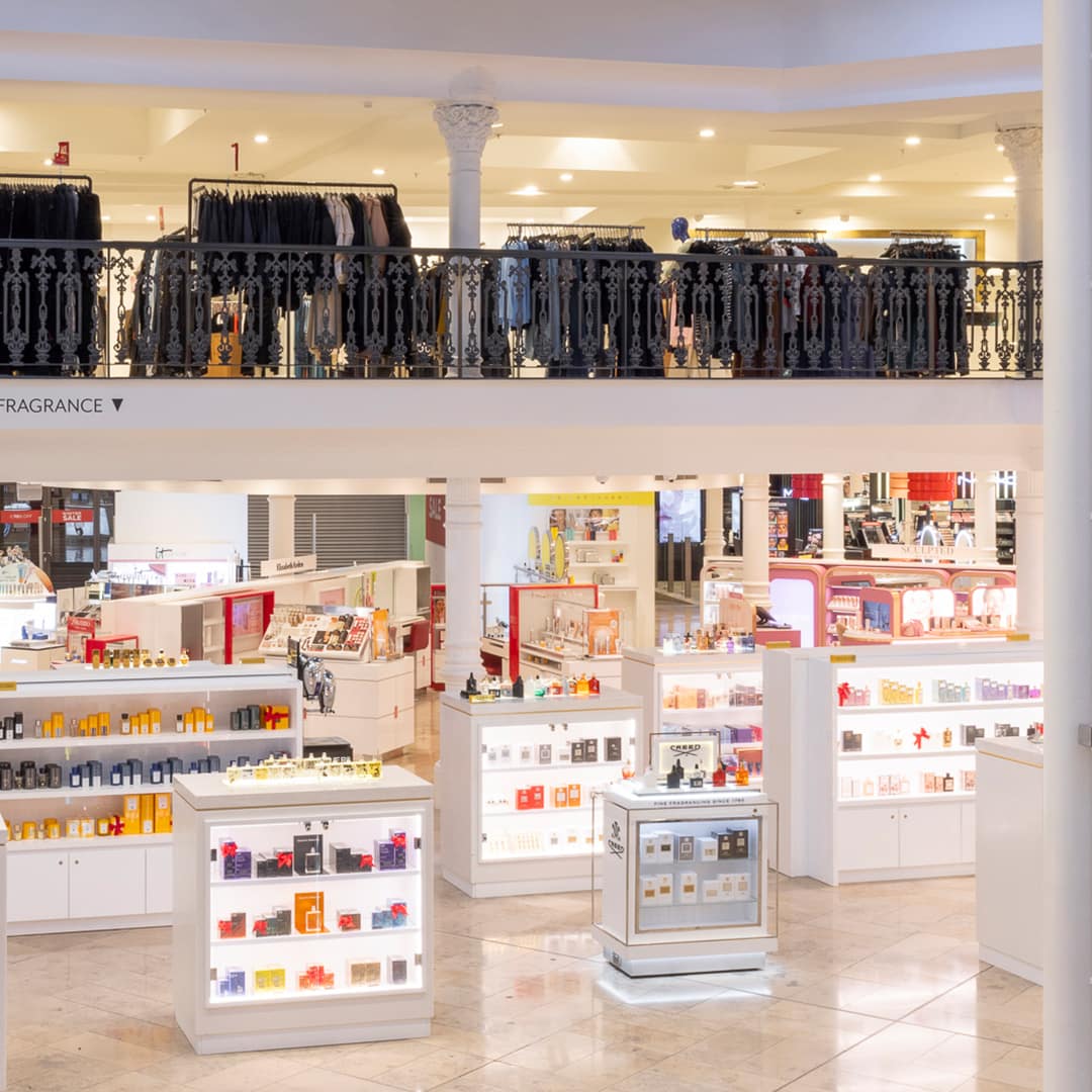 Brightly lit department store interior with a lower level displaying various perfume and cosmetic counters. Shelves are stocked with colourful bottles and products. Upstairs, a black ornate balcony overlooks the floor, with racks of clothes visible.