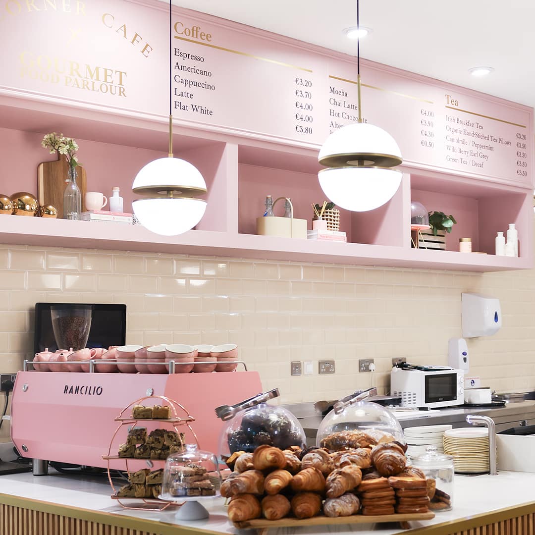 A stylish café interior with pink and cream tones. The counter displays a selection of pastries, including croissants and buns. A pink espresso machine and stacked cups are on the counter. Above, a menu board lists coffee and tea options with prices. Soft pendant lights enhance the cosy atmosphere.