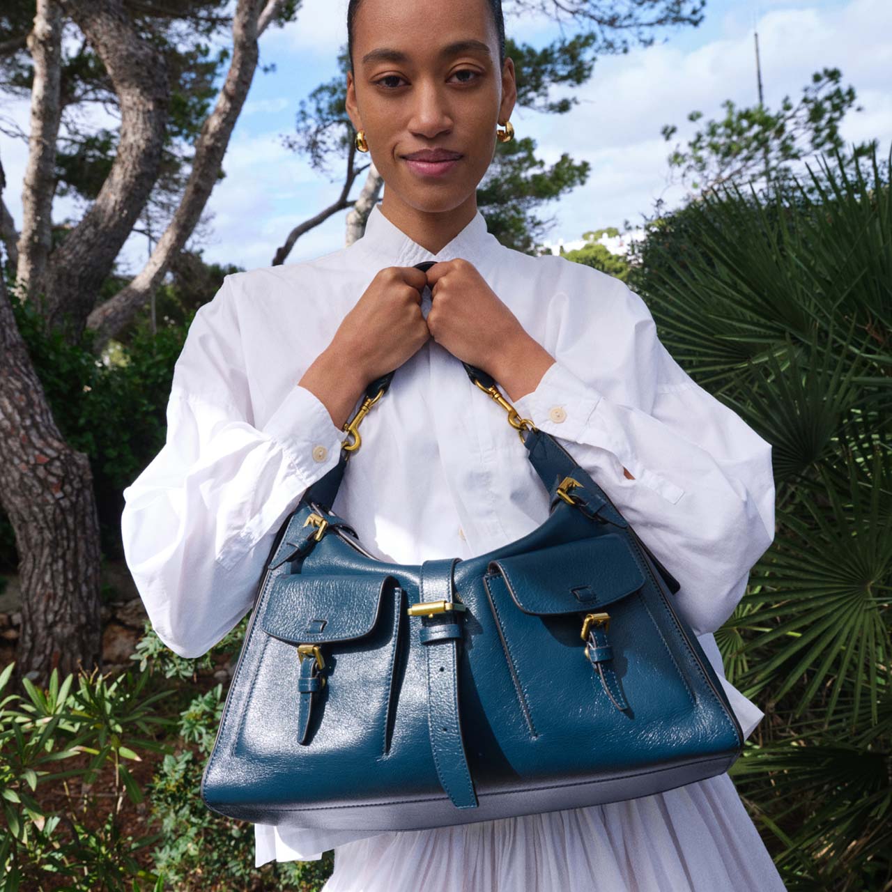 A person holding a blue handbag stands outdoors, surrounded by greenery and trees.