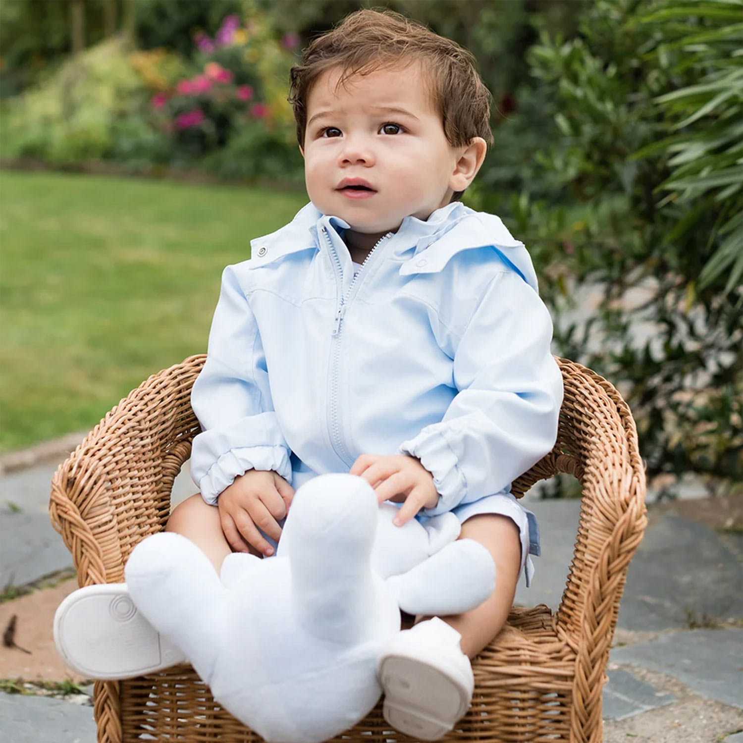 Toddler seated on wicker chair wearing light blue zip-up jacket and white socks, in garden setting