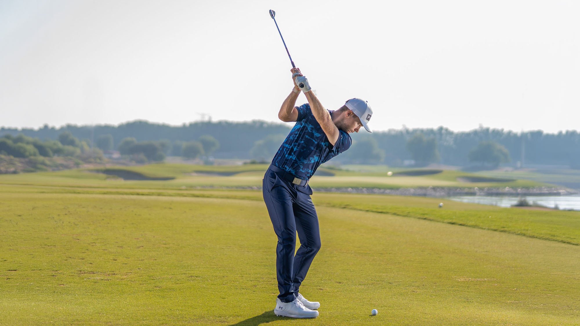 Golfer mid-swing on lush green fairway with coastal landscape and water in background