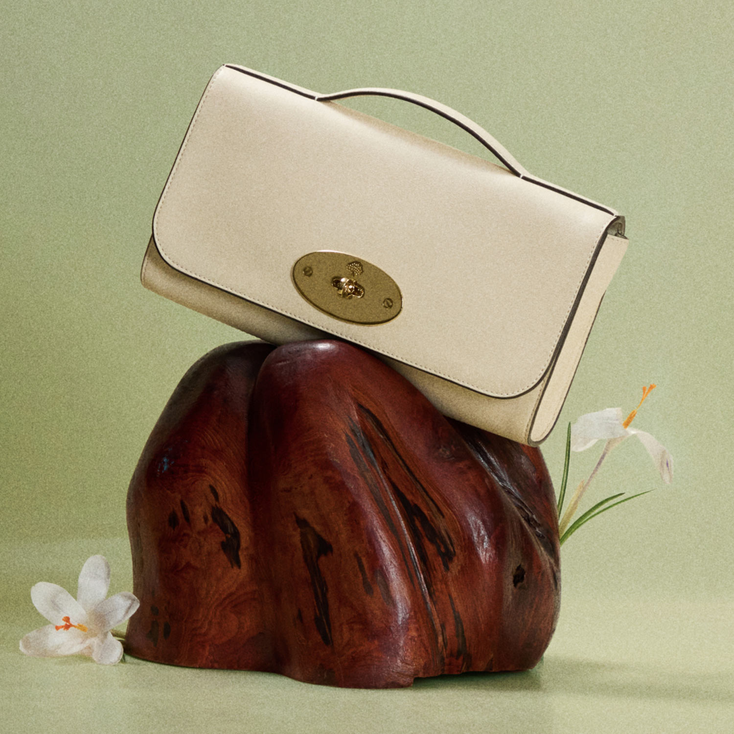 Cream leather shoulder bag with gold clasp displayed on dark wood sculpture with white flowers on mint green background