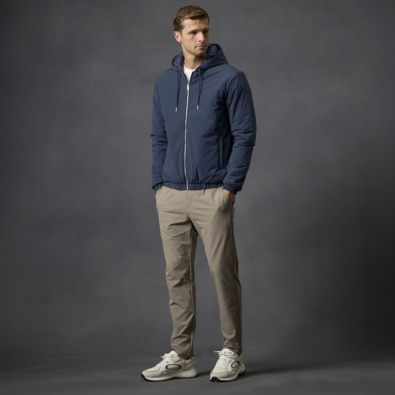 Man wearing navy hooded jacket and beige trousers with white trainers, standing against a grey background.