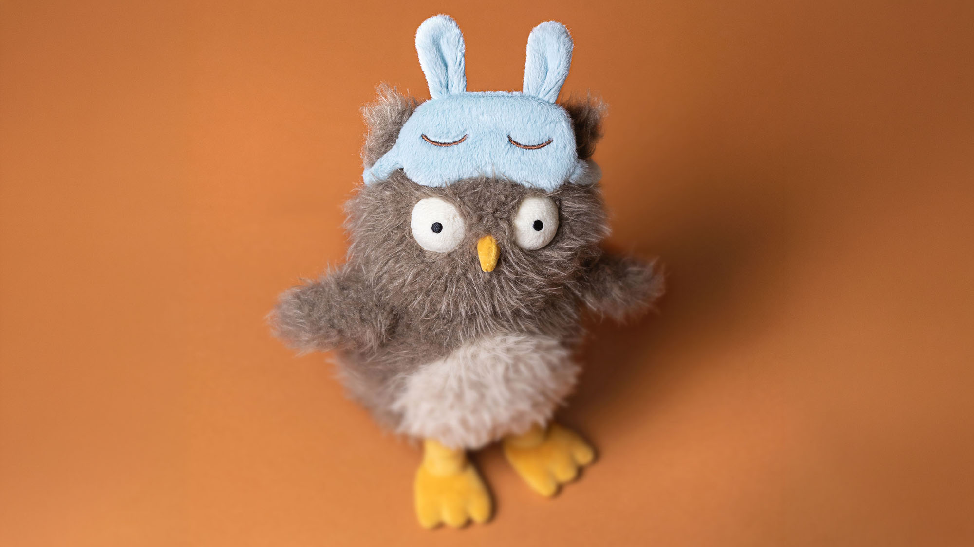 Plush grey owl toy wearing a light blue sleep mask with bunny ears, yellow beak and feet, against an orange background.