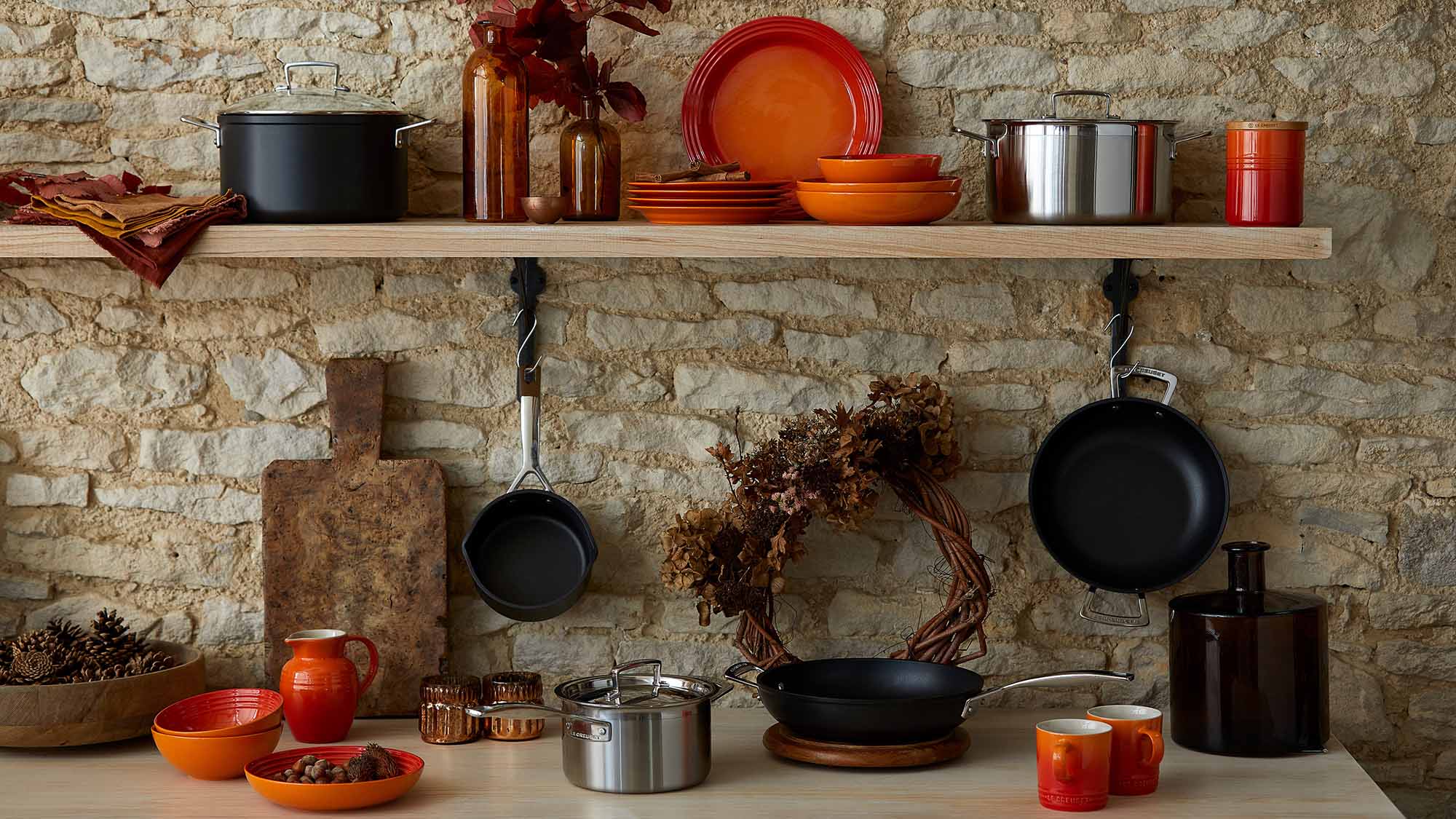 Kitchen shelf with colorful dishes, pots, pans, and autumn-themed decor against a stone wall.
