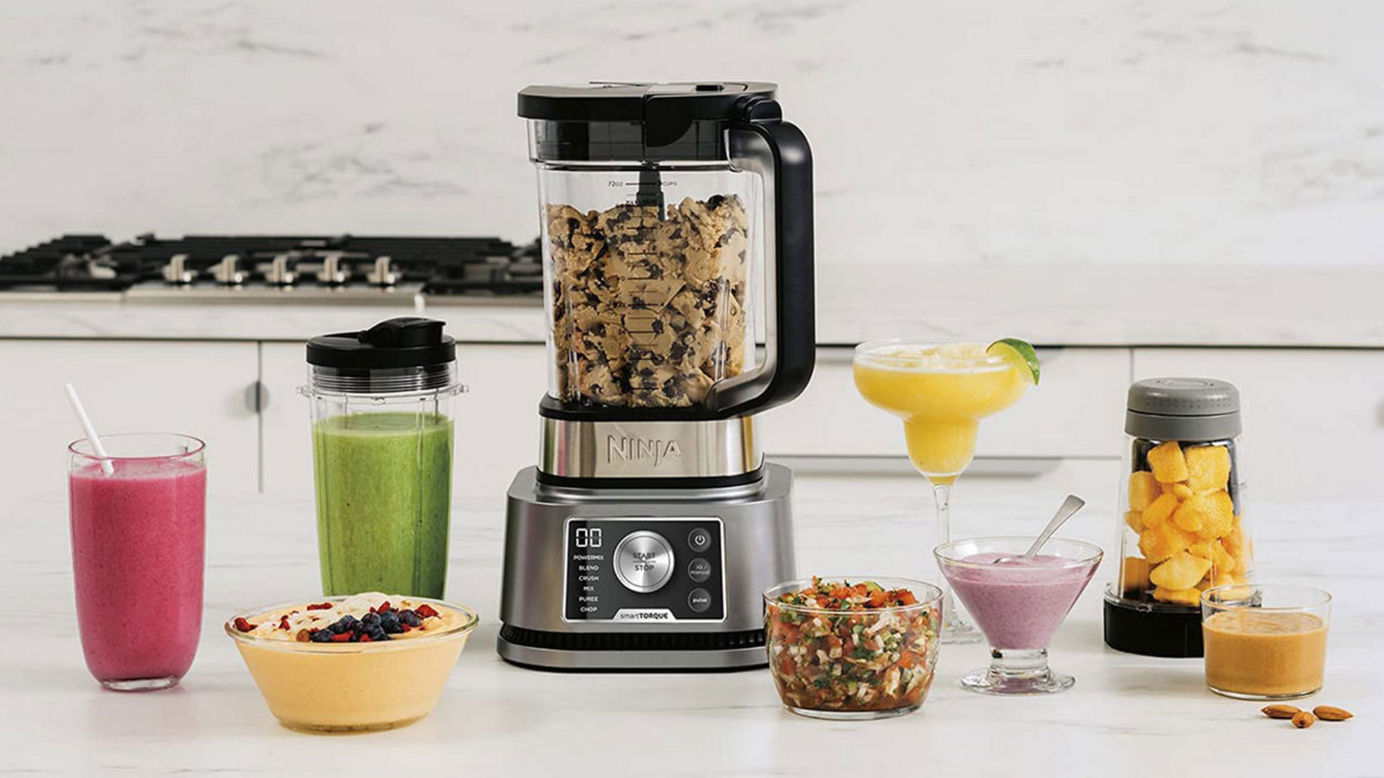 Ninja blender surrounded by smoothies, juices, bowls of food, and fresh ingredients on a kitchen worktop