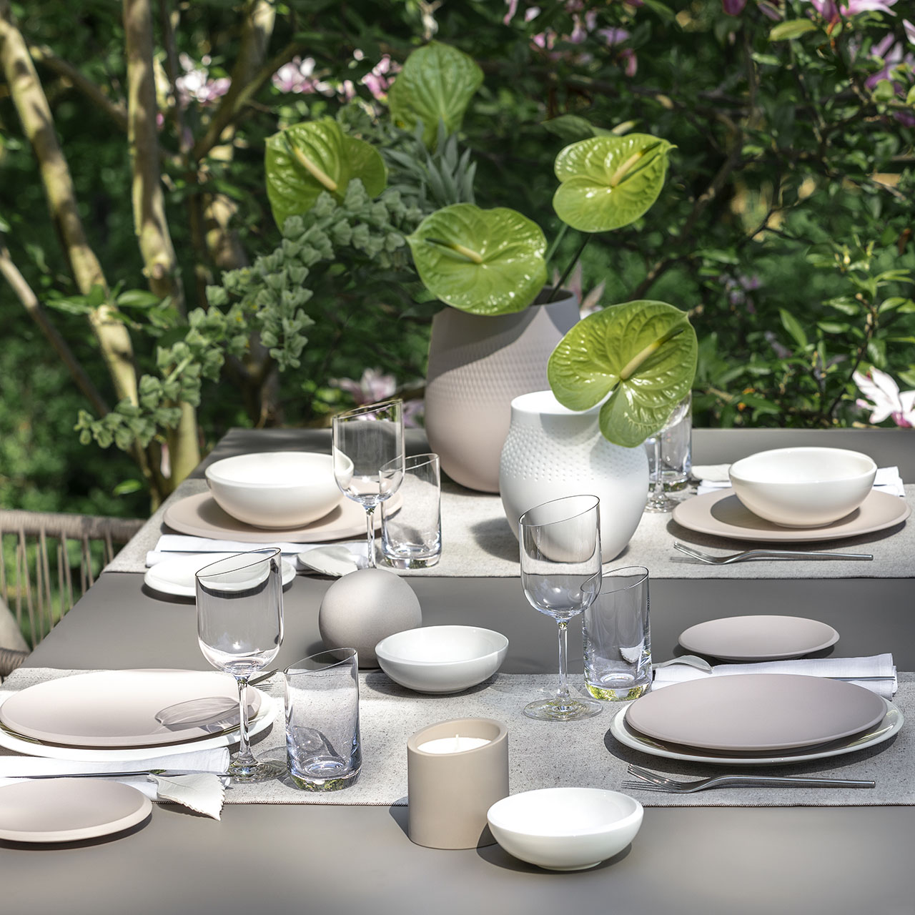 Outdoor dining table set with white plates, glasses, and cutlery, surrounded by green foliage and purple flowers in planters