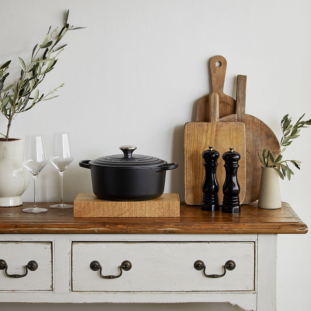 Styled kitchen table with black cast iron cookware, wooden chopping boards, wine glasses, olive branches and pepper mills