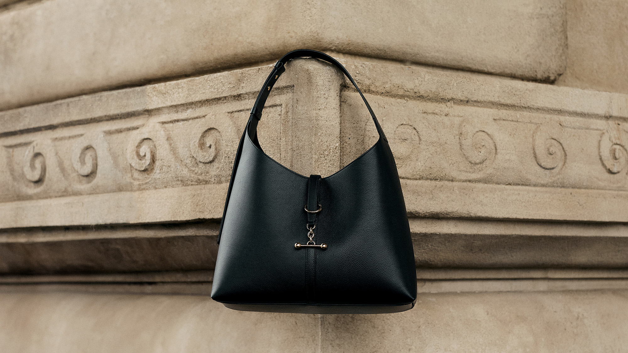 Black leather hobo handbag with single shoulder strap displayed against decorative stone architecture