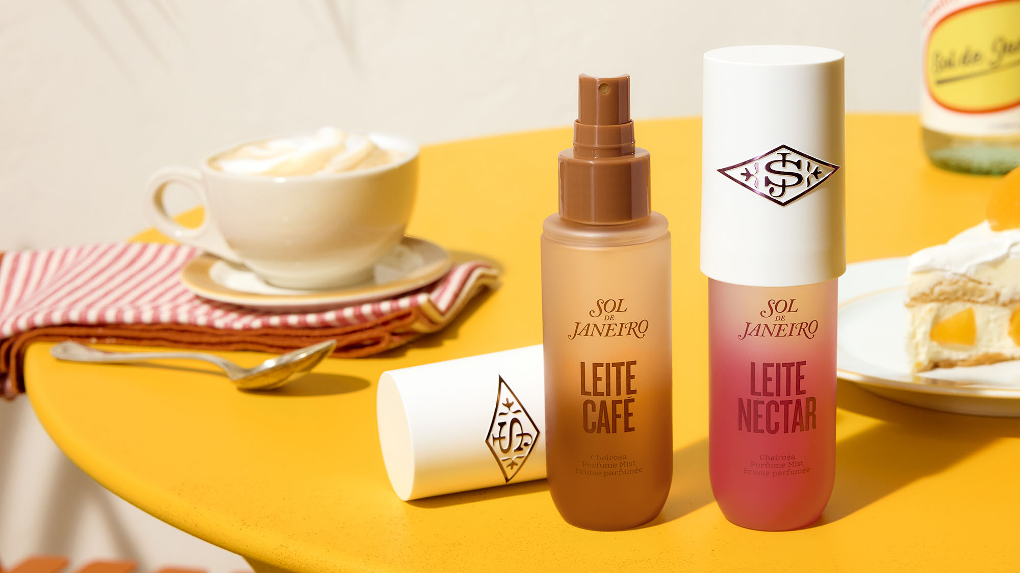 Sol de Janeiro Leite Café and Leite Nectar perfume mists displayed on yellow surface with coffee cup and cake