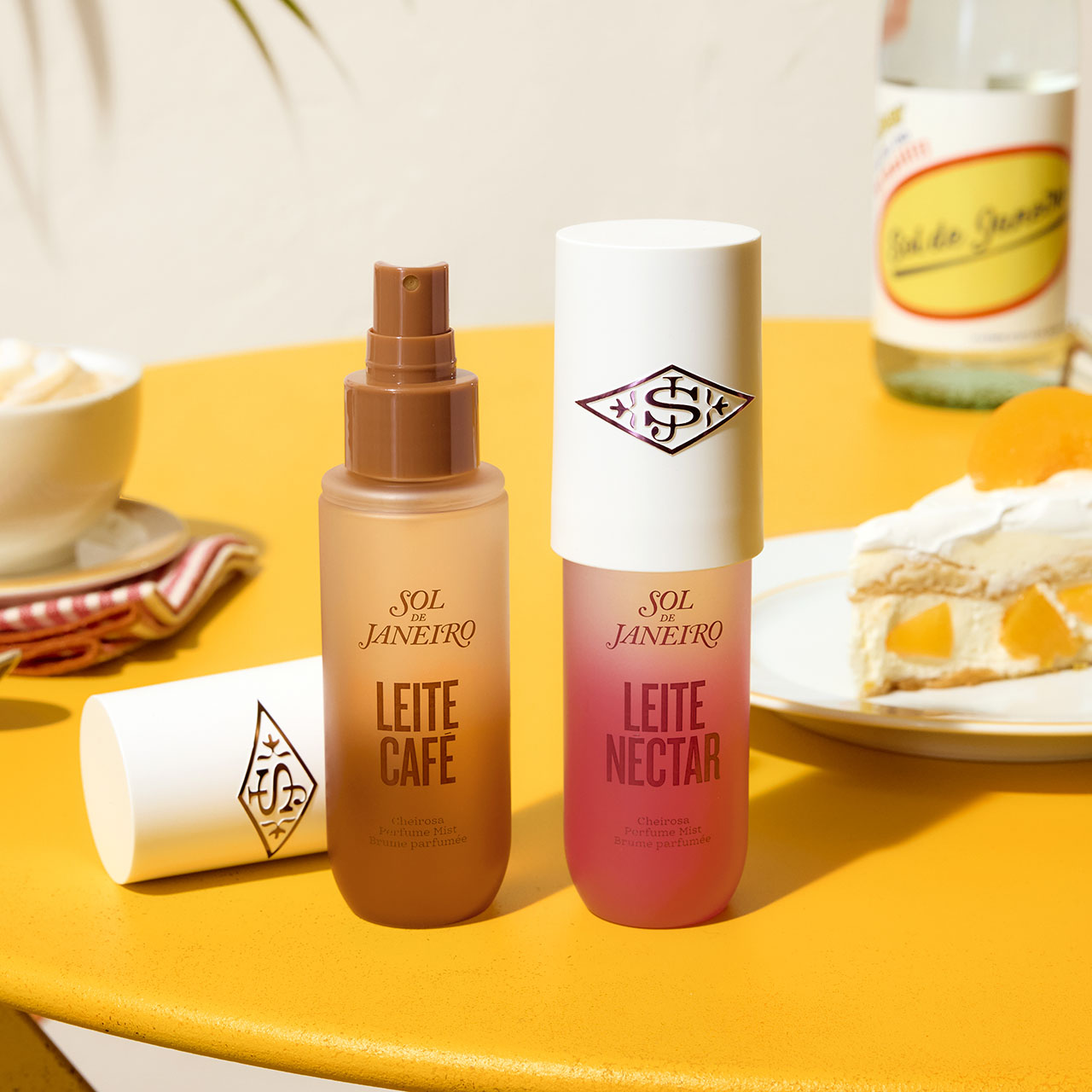Sol de Janeiro Leite Caf&eacute; and Leite Nectar perfume mists displayed on yellow surface with coffee cup and cake