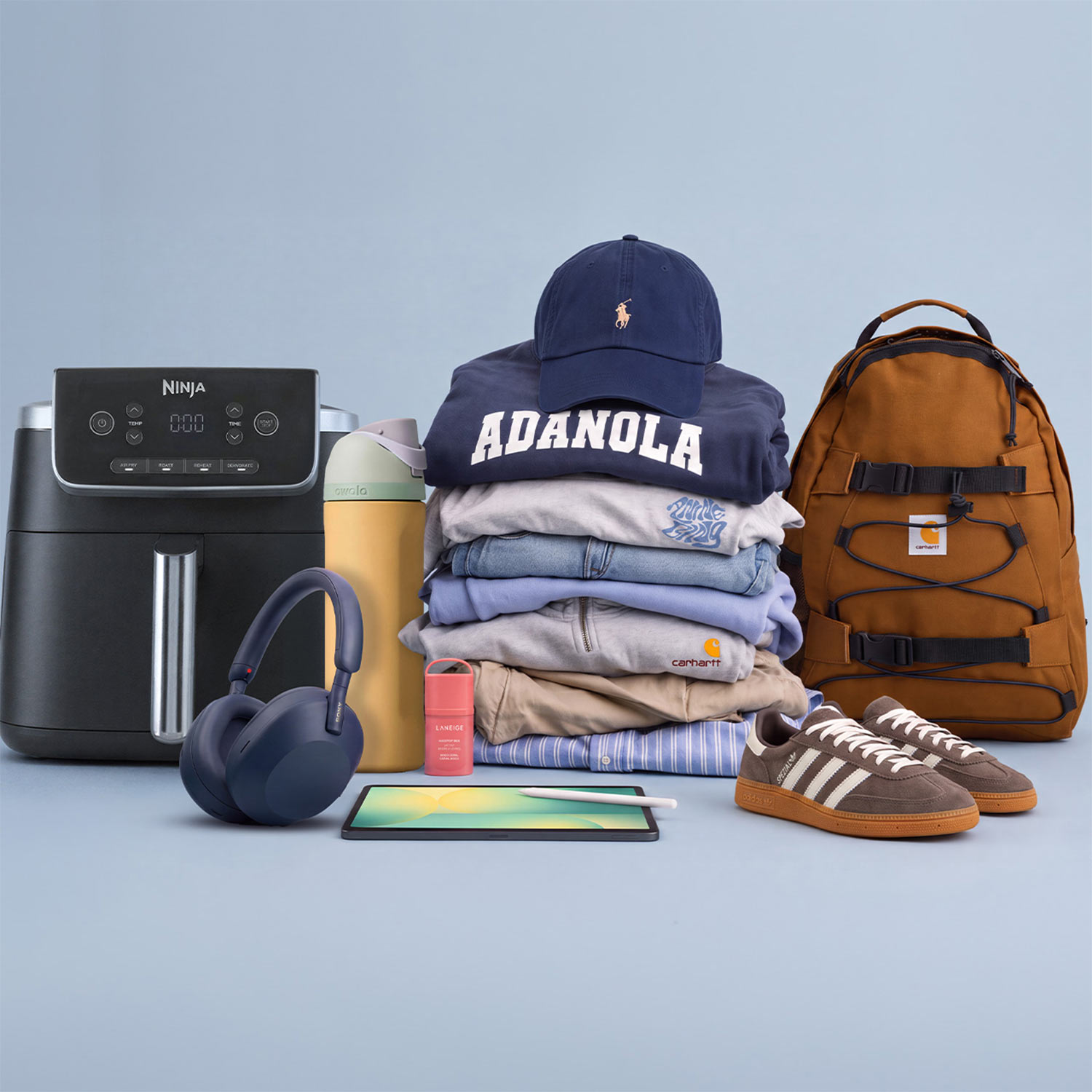 Curated collection of student essentials including air fryer, wireless headphones, yellow water bottle, navy baseball cap with logo, stack of folded clothing, brown canvas backpack, tablet, trainers and setting spray.