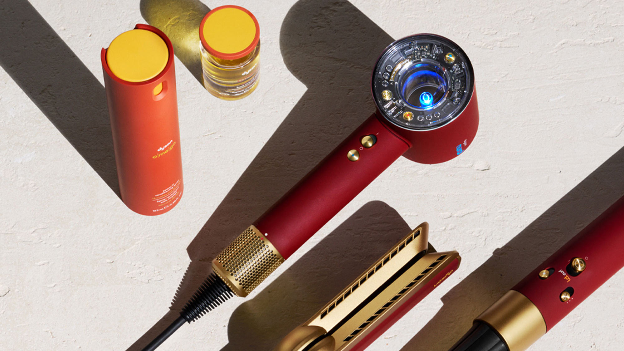 Collection of Dyson hair styling tools including red cylindrical devices, hair dryer with blue display, round brush, and straightener on white surface