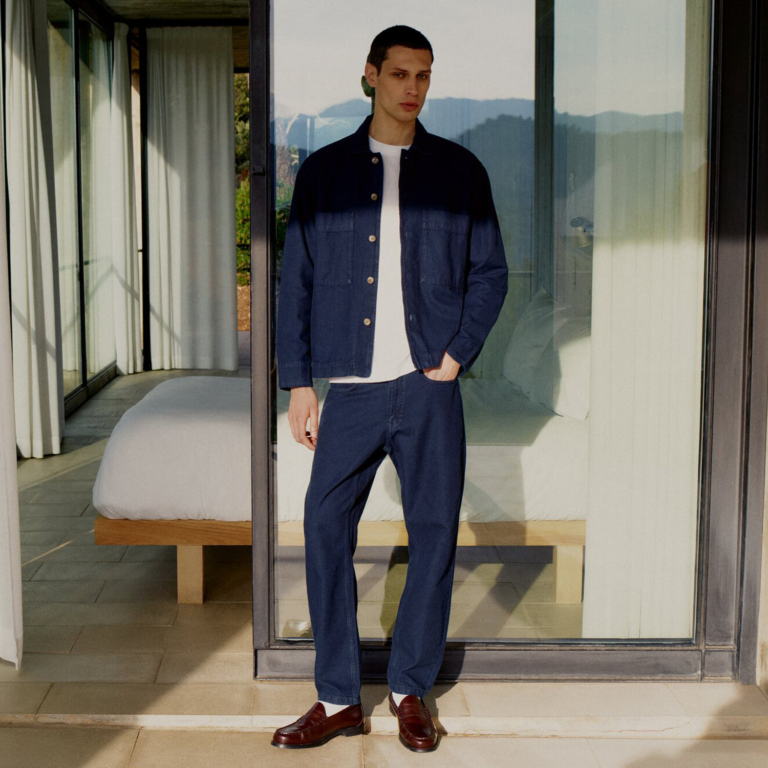 Man wearing navy colour-block denim jacket and matching trousers with burgundy loafers, standing in modern interior space