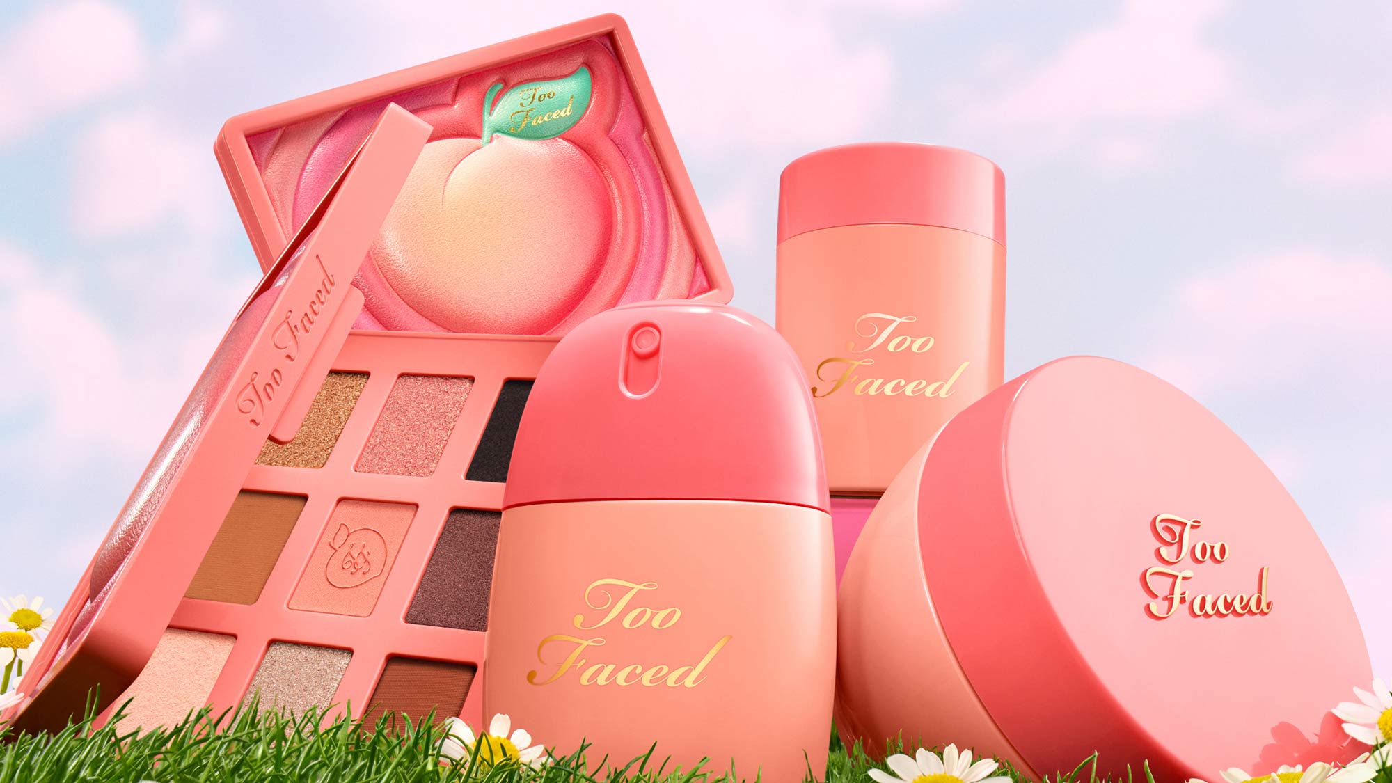 Too Faced cosmetics collection featuring pink eyeshadow palette, liquid highlighter, and powder products arranged on grass with daisies