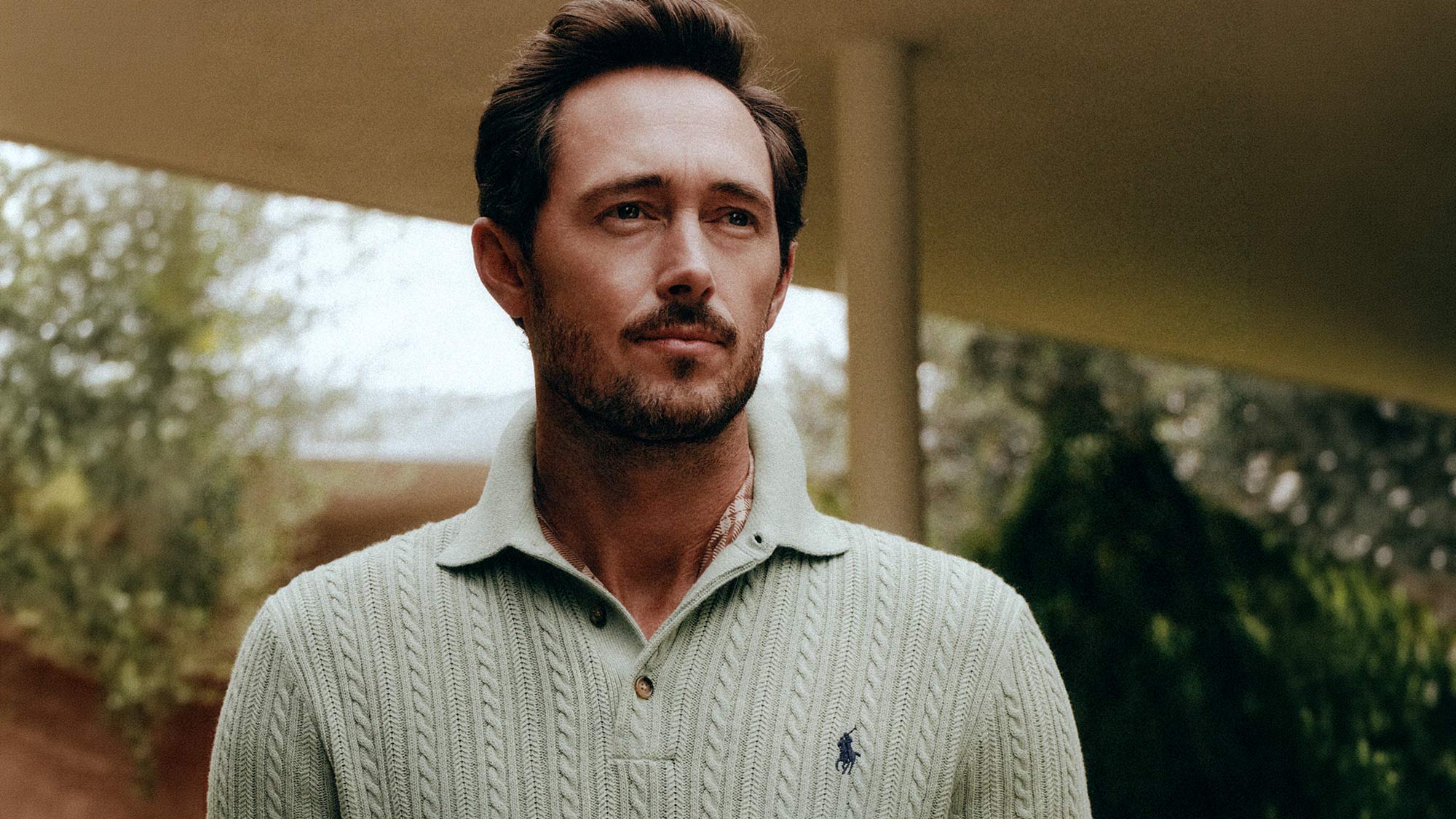Man with dark hair and beard wearing a cream cable-knit polo shirt with embroidered logo, standing outdoors with greenery in background.