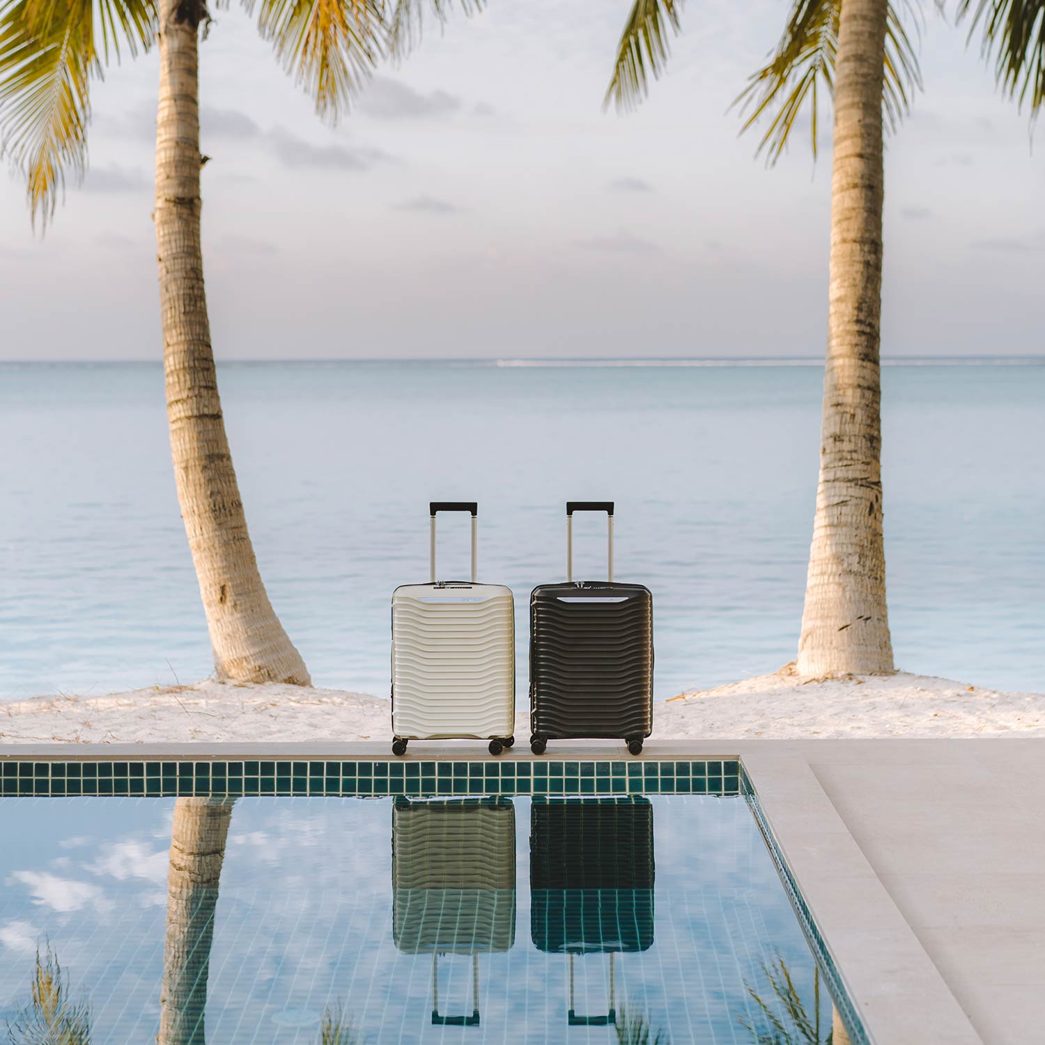 Cream and black rolling suitcases positioned between palm trees overlooking tropical beach and pool