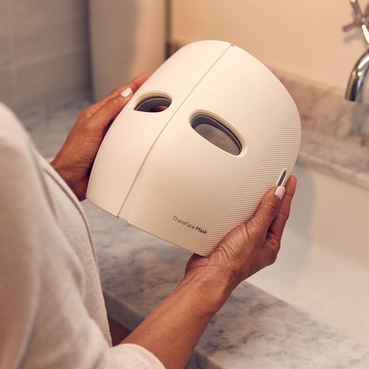 Woman holding white TheraFace Mask device with oval eye openings for facial treatment