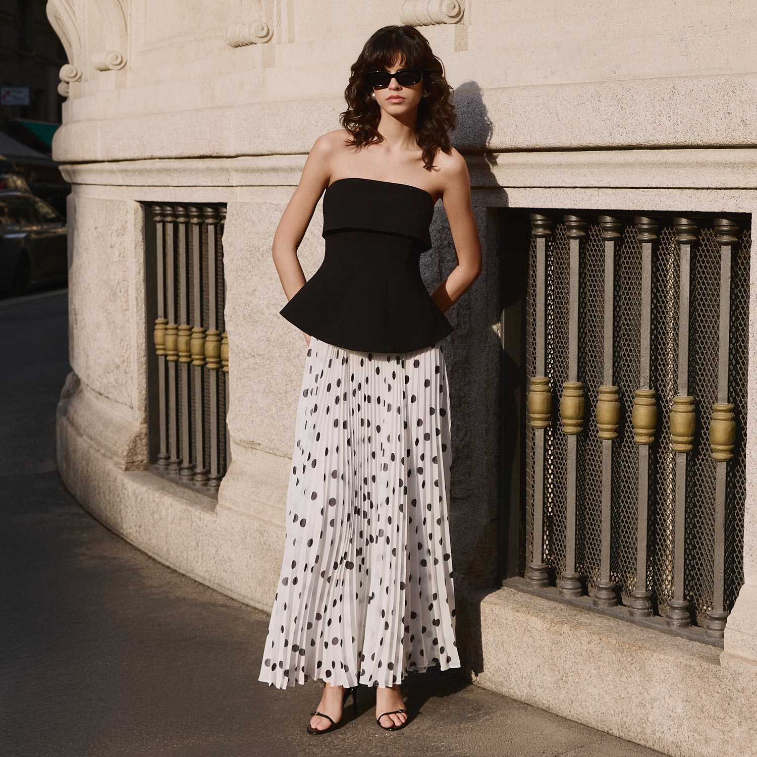 Woman wearing black strapless top with white polka dot pleated midi skirt and sunglasses, standing against stone building
