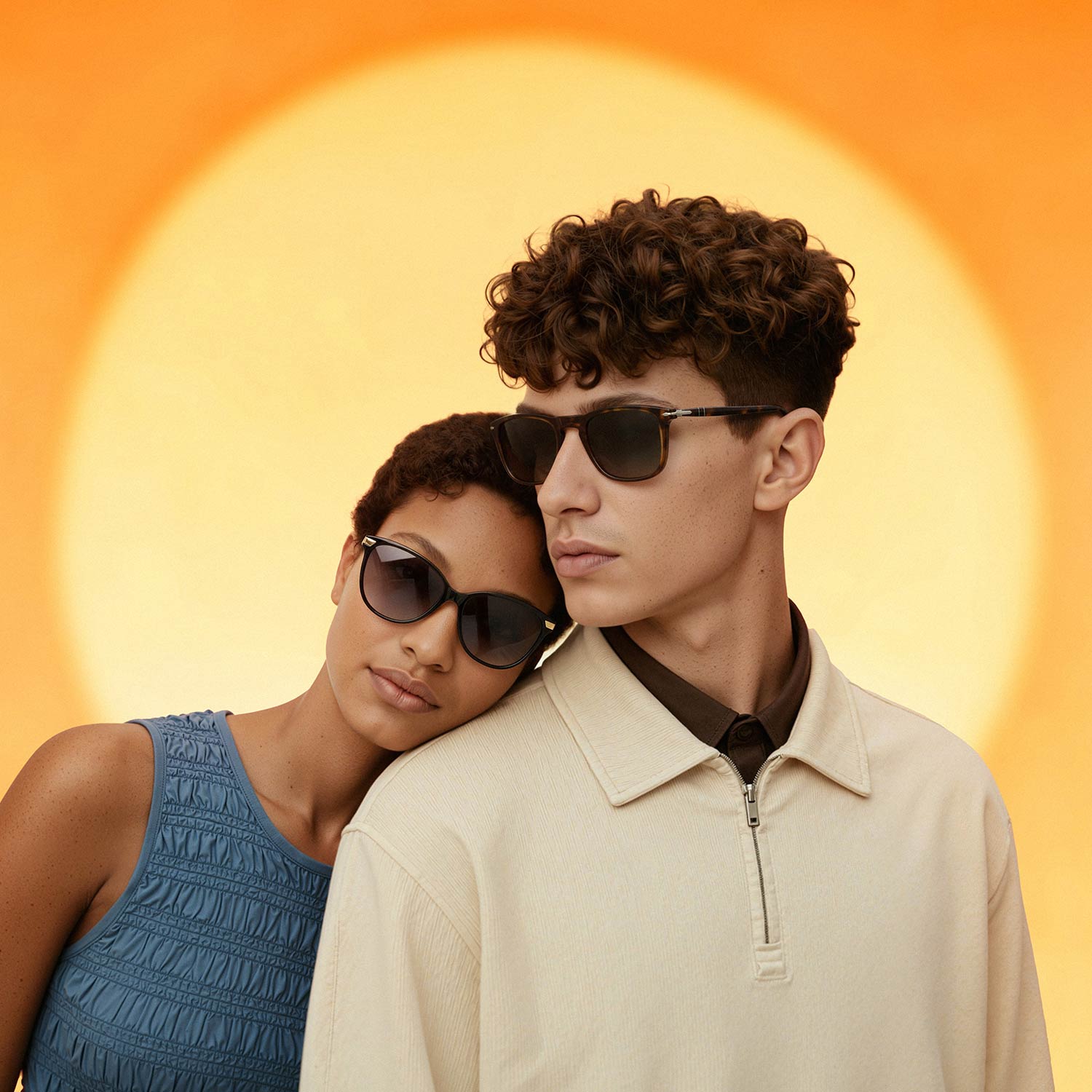 Two people wearing dark-tinted sunglasses against a warm yellow background. Woman in blue textured top, man in cream zip-neck jumper.