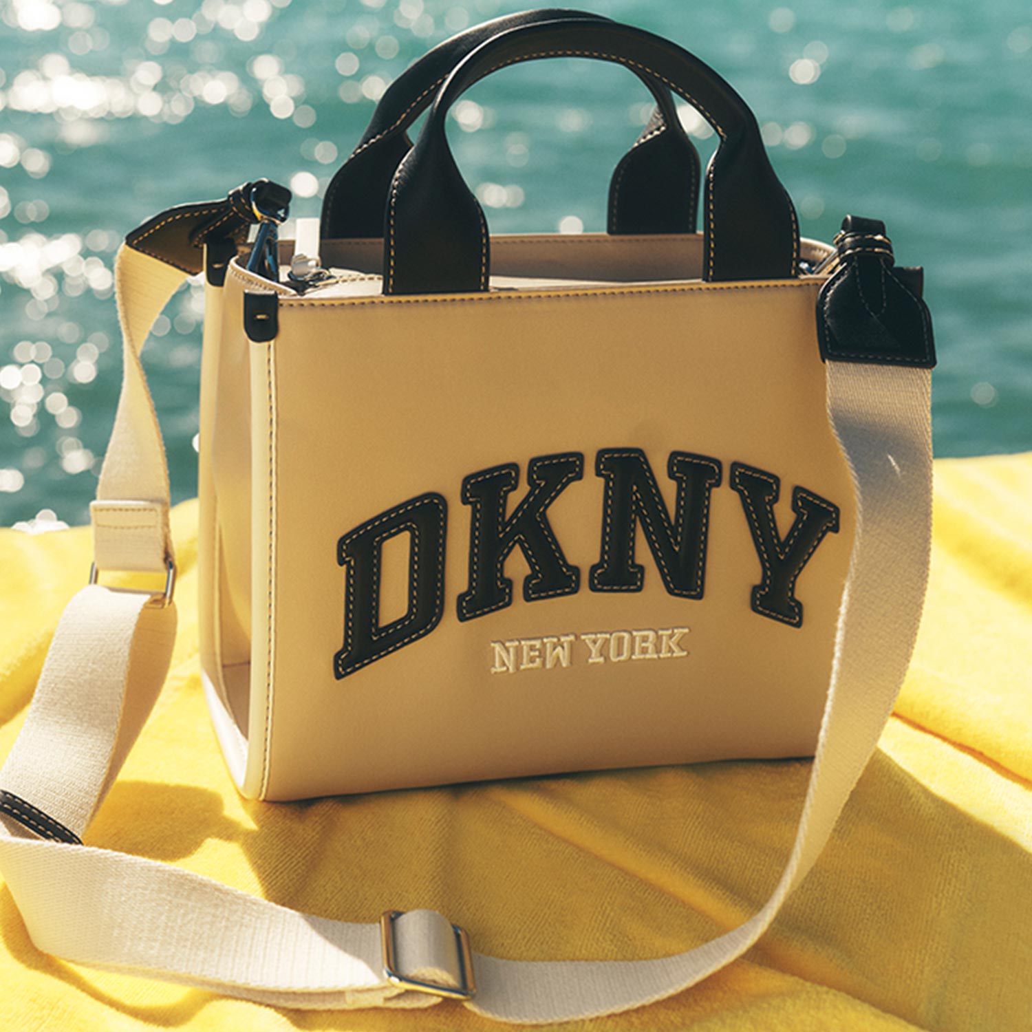 Cream tote bag with black DKNY New York branding and black handles, displayed on yellow fabric by turquoise sea.