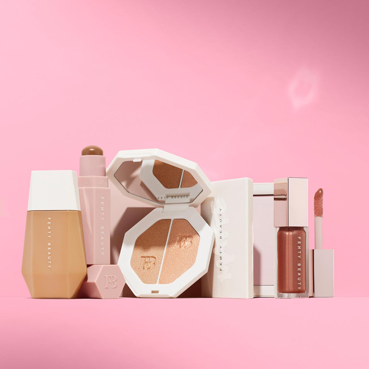 Fenty Beauty cosmetics collection including foundation, highlighter, lip oil and liquid lipstick in neutral and warm tones on pink background