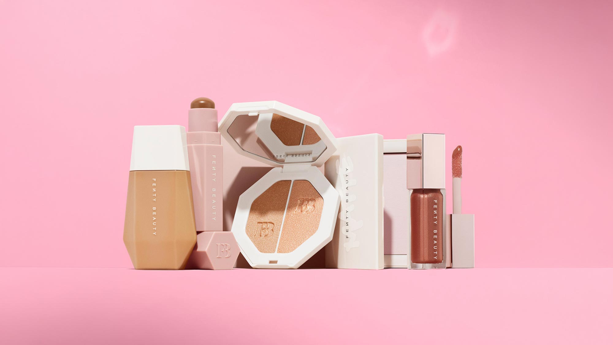 Fenty Beauty cosmetics collection including foundation, highlighter, lip oil and liquid lipstick in neutral and warm tones on pink background