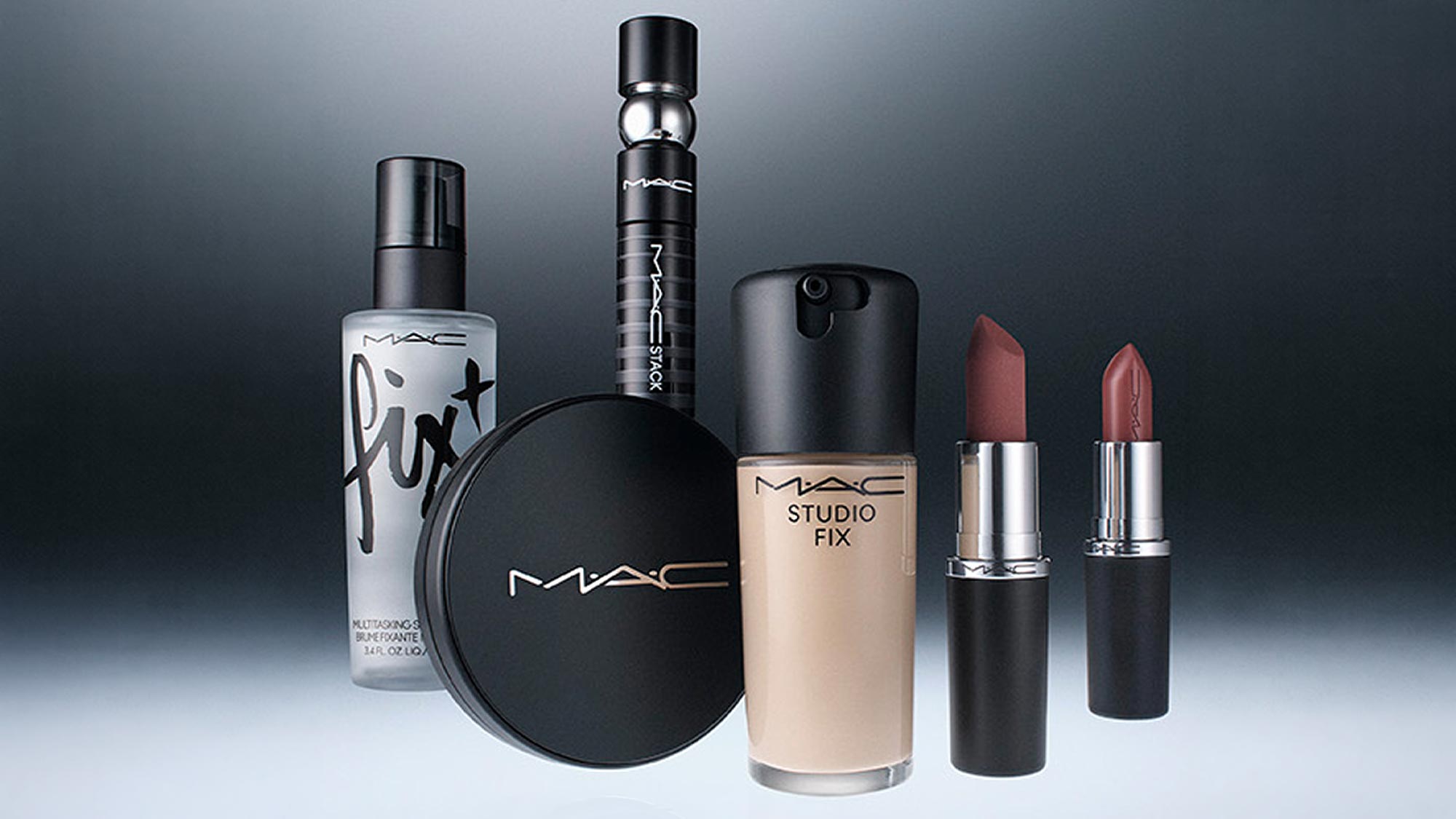 M.A.C cosmetics collection including foundation, lipsticks, setting spray, and compact powder on grey background