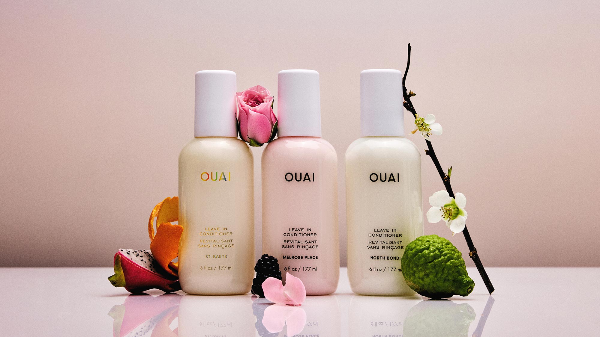 Three OUAI Leave In Conditioner bottles in Melrose Place, St. Barts and North Bondi scents, surrounded by fresh flowers and citrus fruits on pink background.
