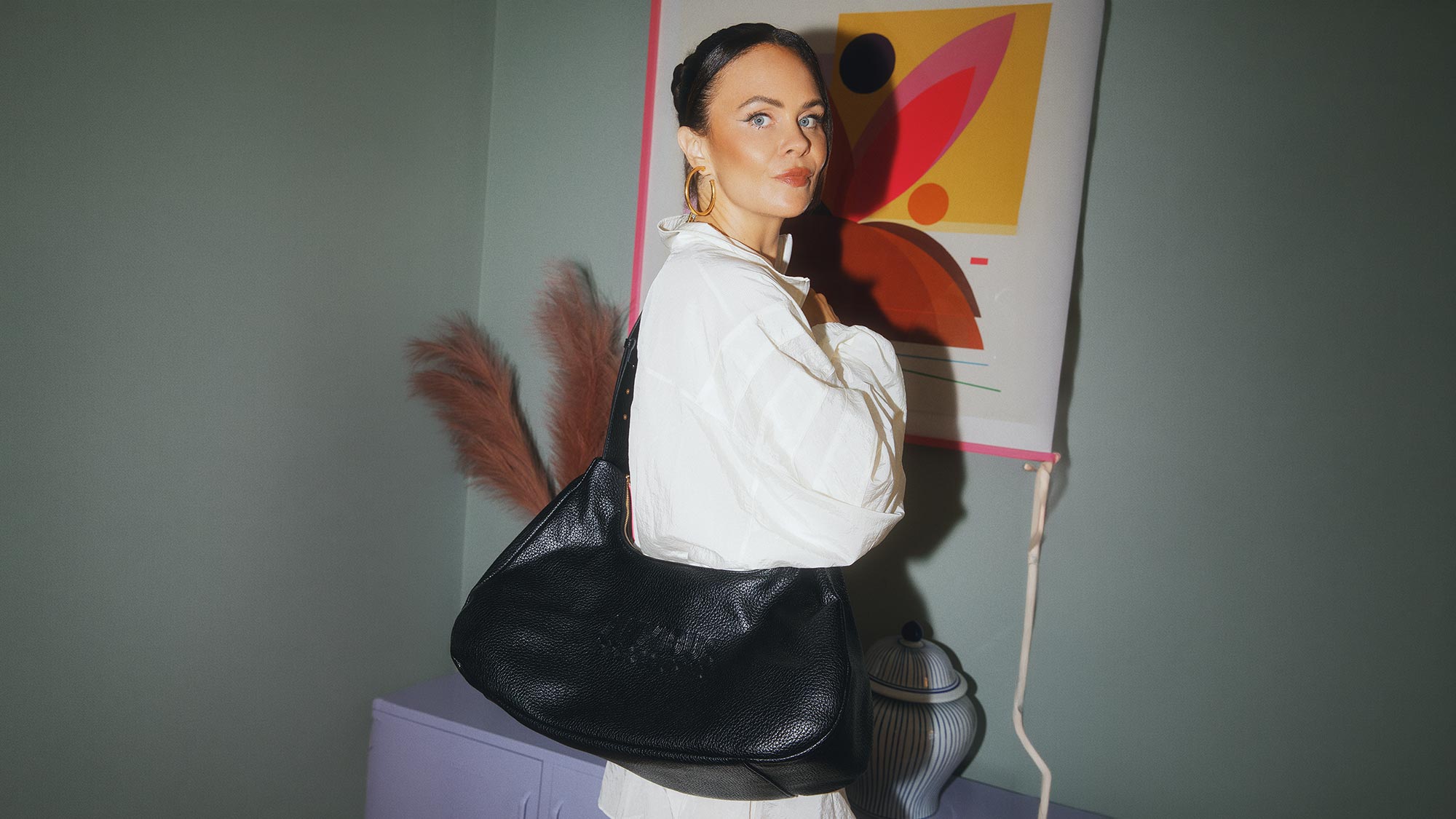 Woman wearing white shirt and gold hoop earrings carrying black textured shoulder bag next to colourful abstract artwork