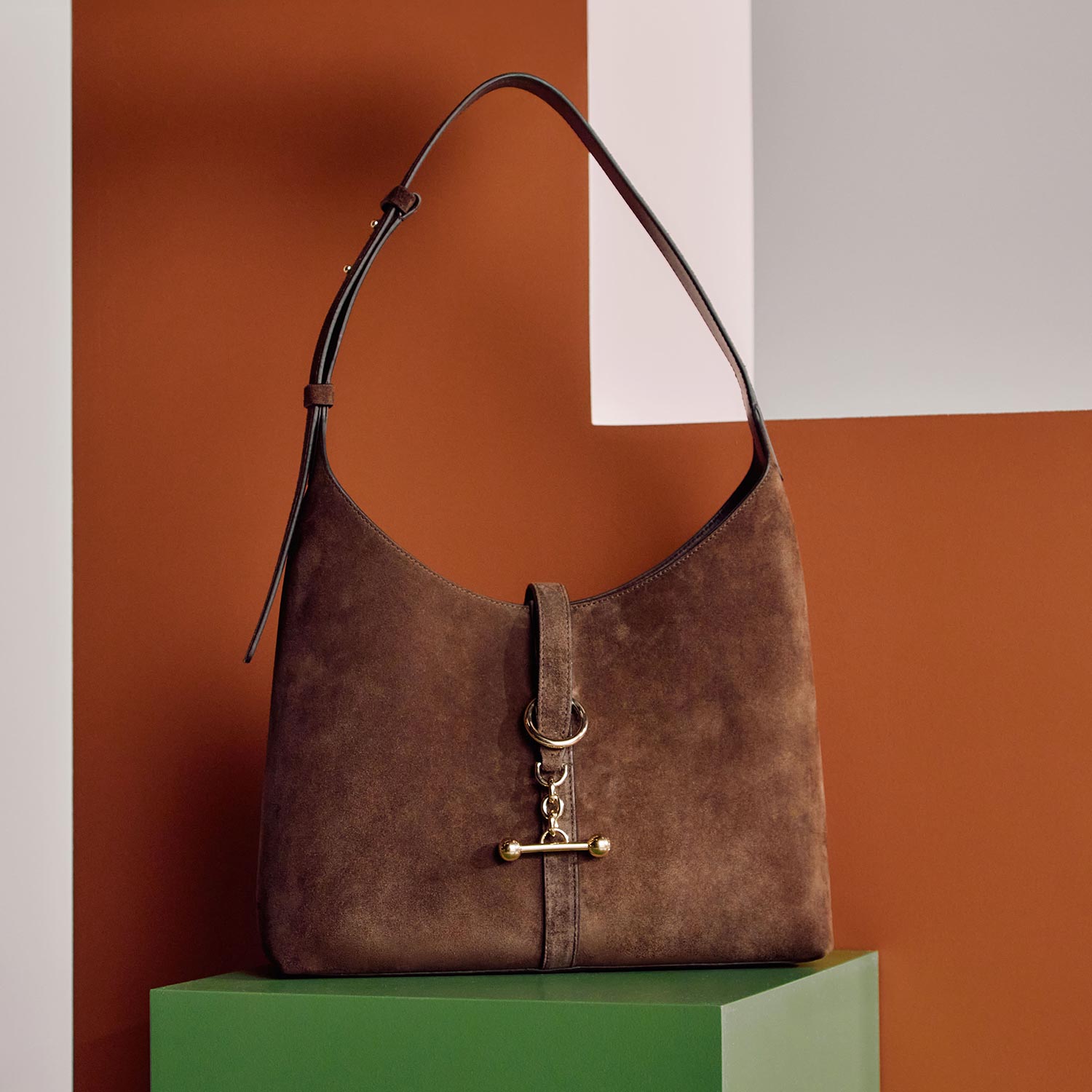 Brown suede shoulder bag with leather strap and metal toggle detail displayed on green platform against orange backdrop