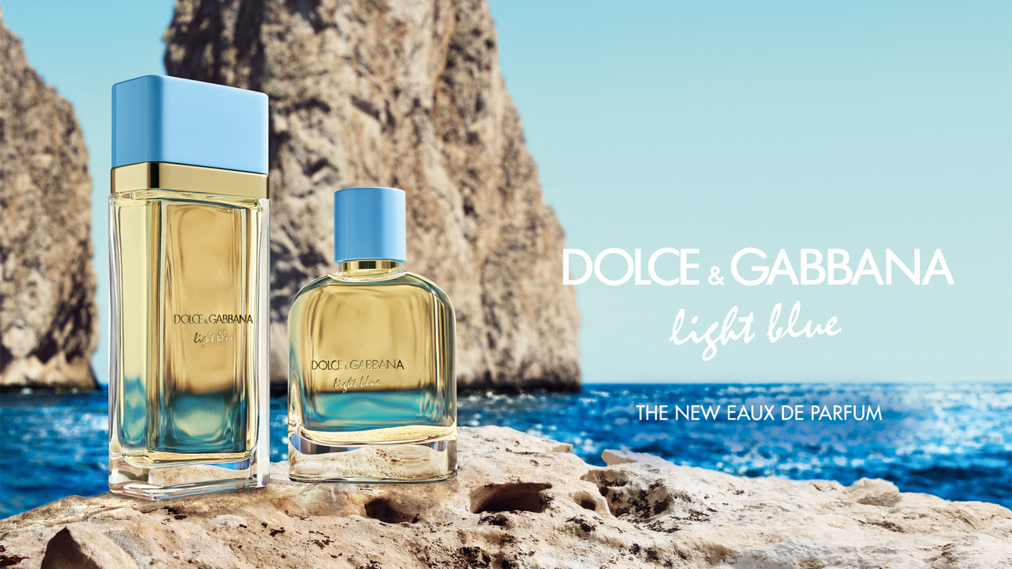 Two Dolce & Gabbana Light Blue perfume bottles on a rocky shore with sea and cliffs in the background.