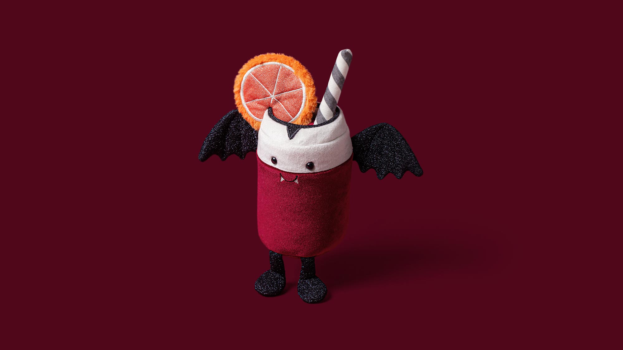 Playful plush vampire character with red body, black wings and sparkly details, holding a grapefruit slice against burgundy background