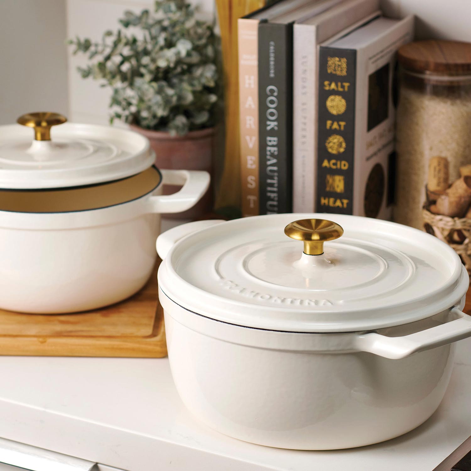 White and cream enamelled cast iron cookware with brass knobs displayed on wooden board with cookbooks and plant