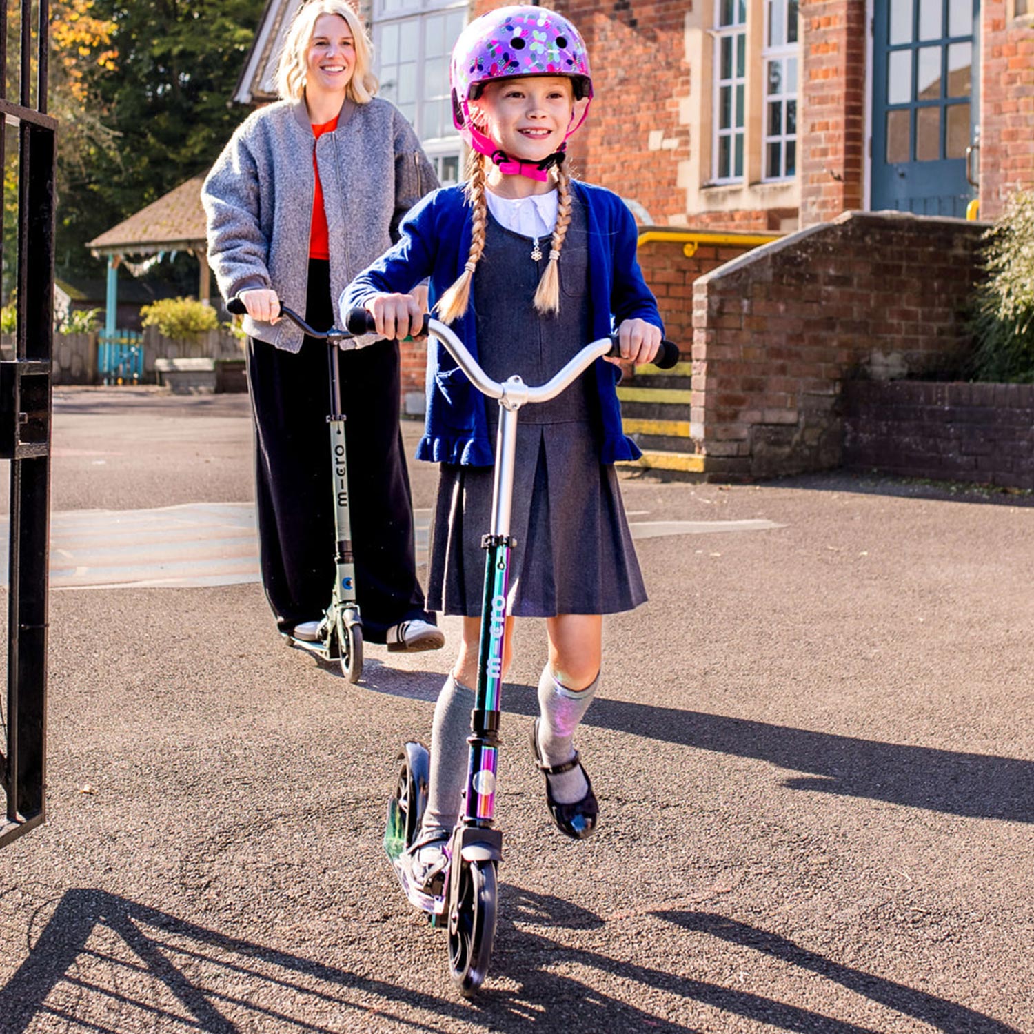Child in pink helmet riding rainbow-coloured Micro scooter with adult on scooter behind in brick courtyard setting