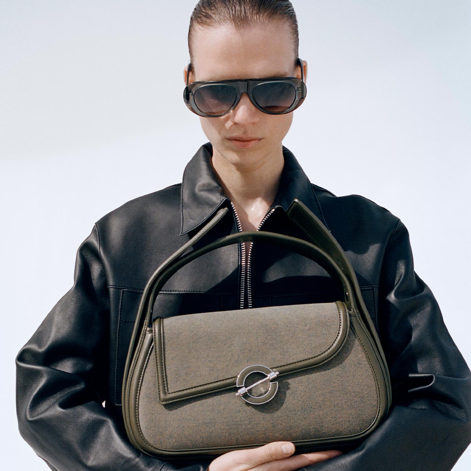 Woman wearing black leather jacket holding taupe and olive handbag with circle ring detail