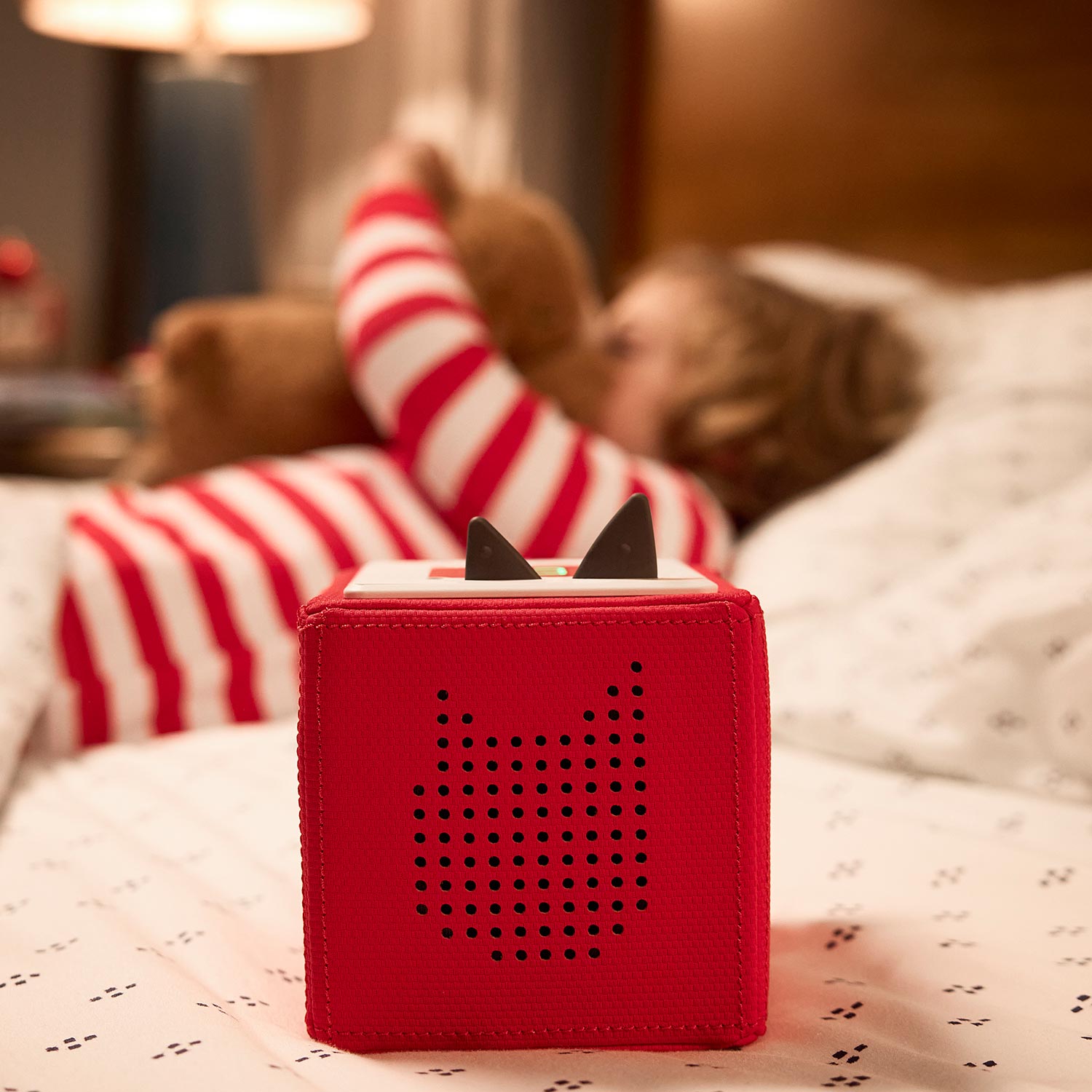 Red Tonie audio player with cat ears on bed with child wearing red and white striped socks in background