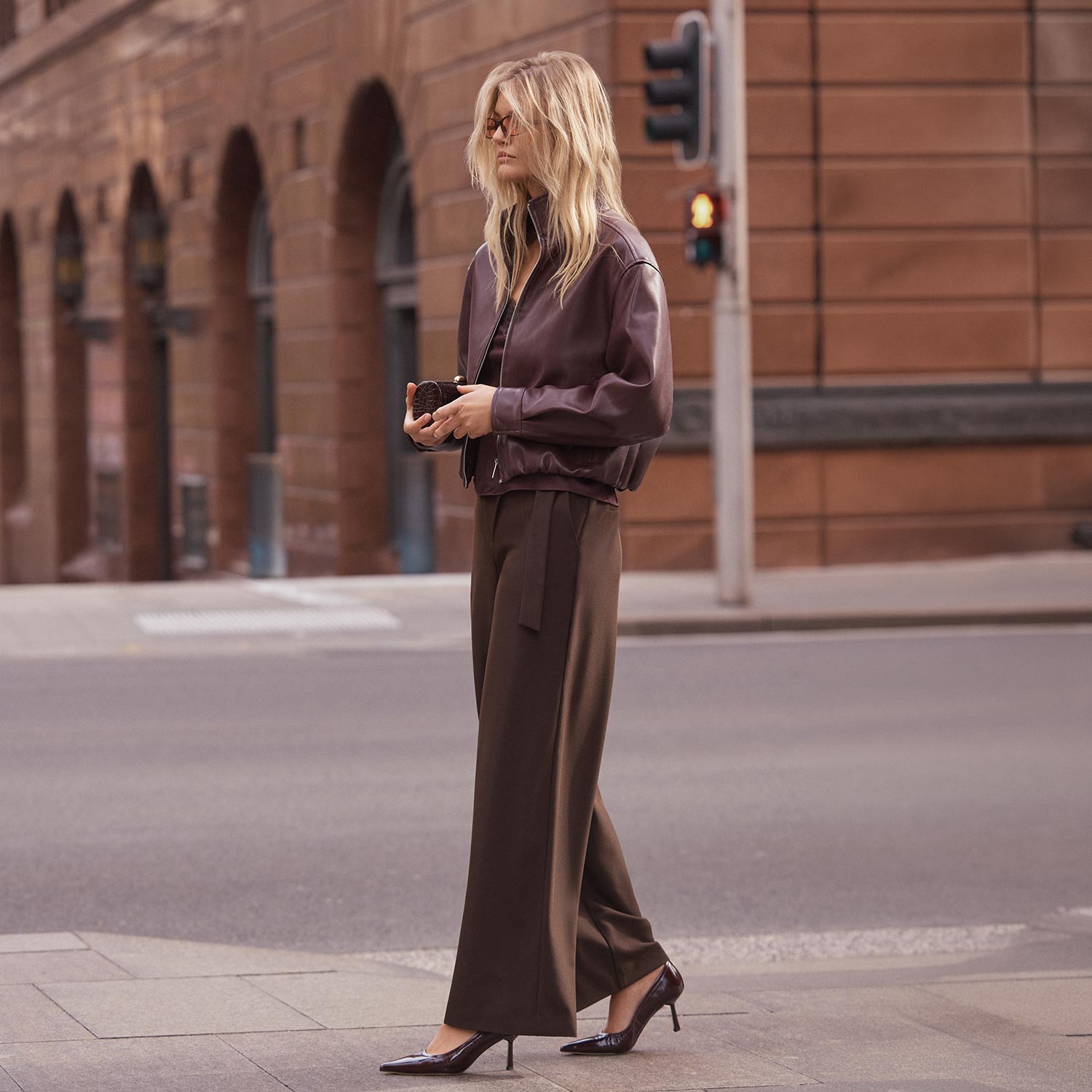 Woman in purple bomber jacket and olive trousers standing on urban street holding small bag