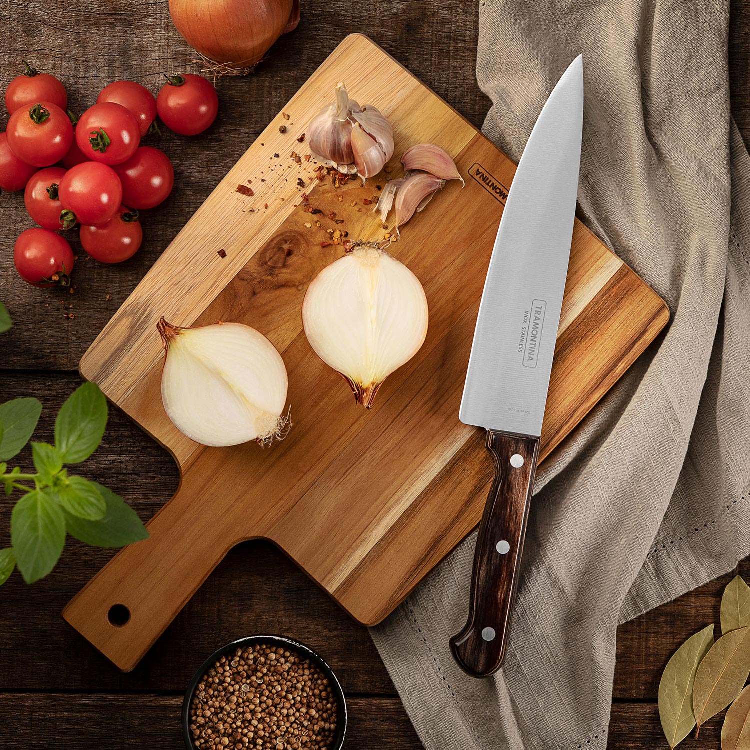 Wooden chopping board with sliced onions, garlic, tomatoes, fresh herbs, and a chef's knife with brown handle on dark wood surface