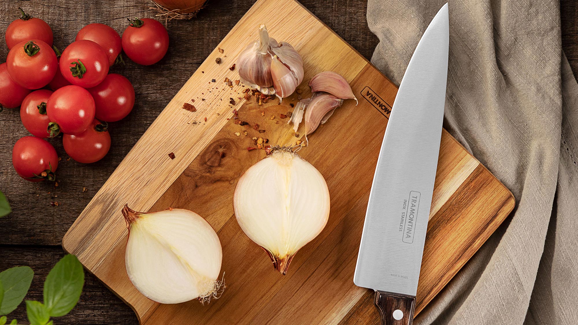 Wooden chopping board with sliced onions and garlic, chef's knife, fresh tomatoes and linen cloth on dark wood surface