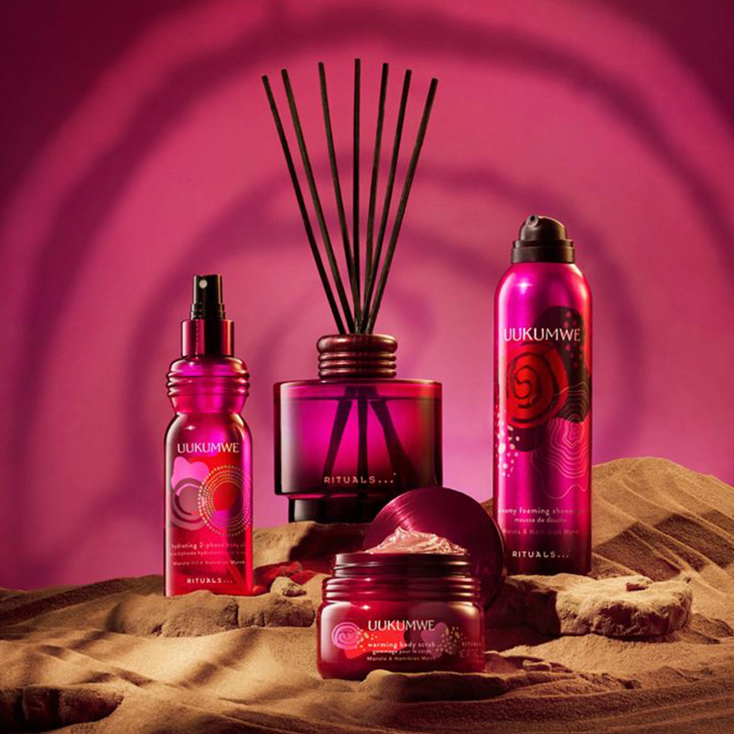 Rituals Uukumwe collection beauty products in deep red bottles and diffuser arranged on sandy surface against pink background