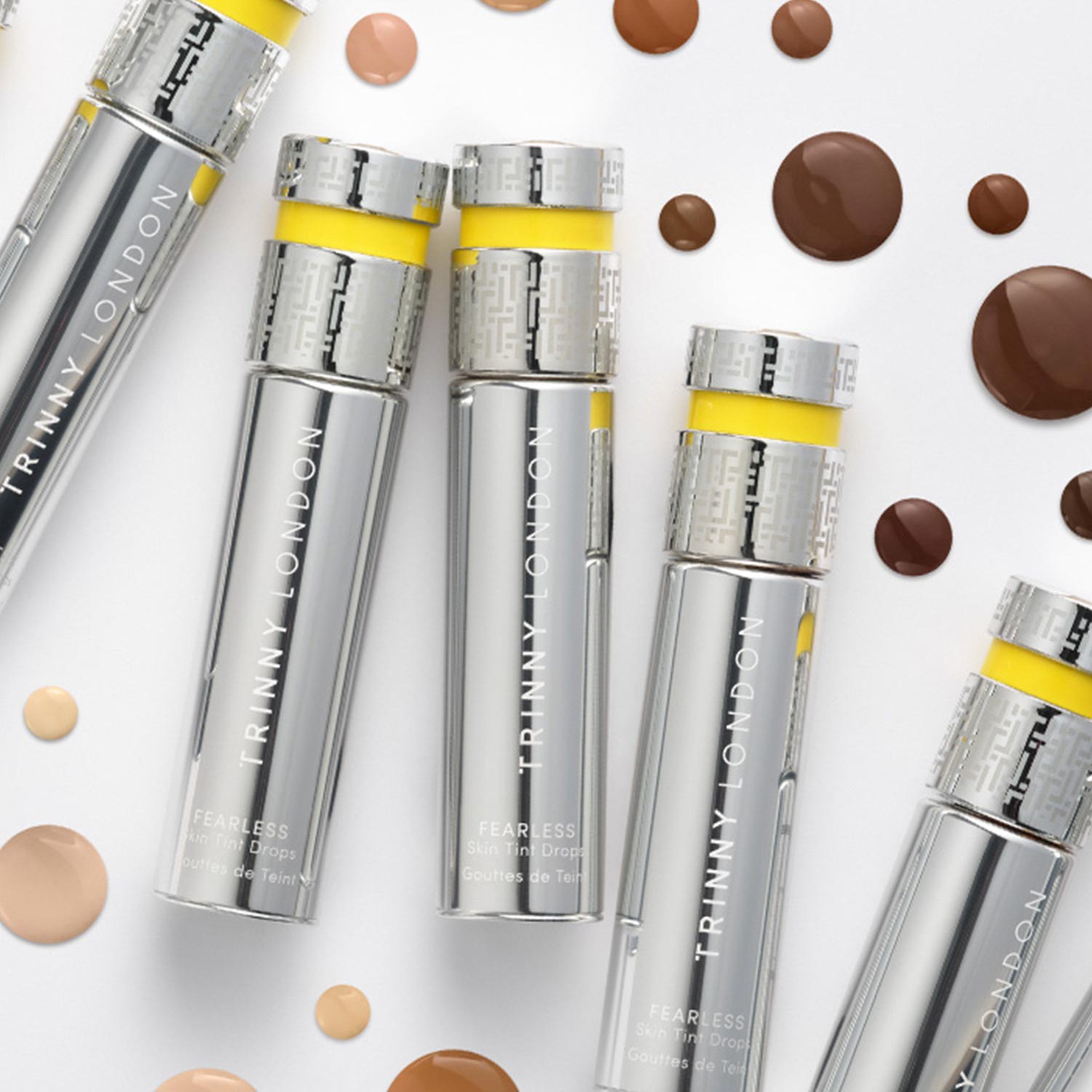 Trinny London Fearless Skin Tint Drops bottles with yellow accents surrounded by swatches of foundation shades