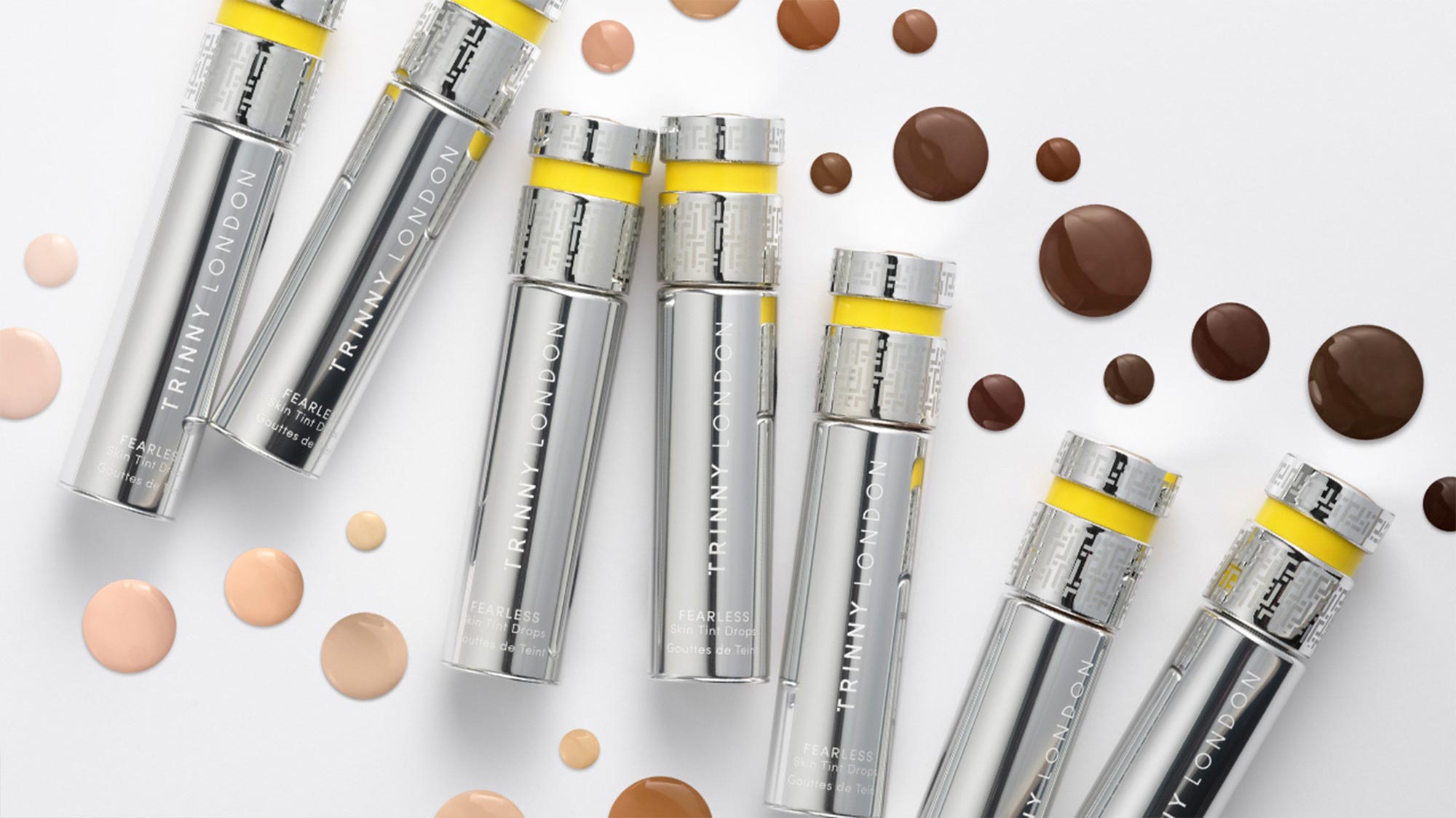 Trinny London Fearless Skin Tint Drops bottles with silver and yellow accents surrounded by foundation swatches in various skin tones
